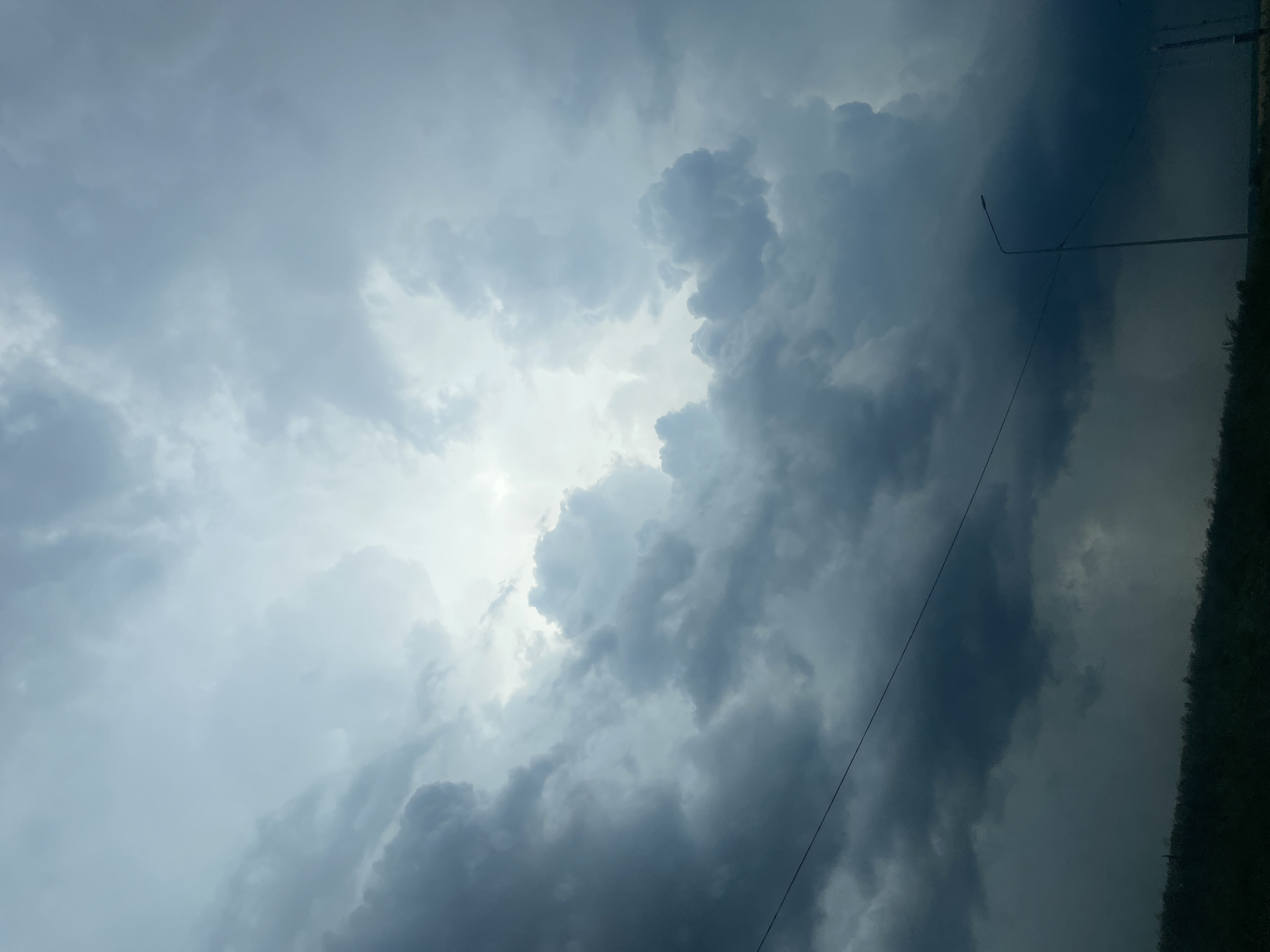This is a low-angle, full shot of dark, ominous storm clouds gathering in the sky over the city of Unknown, Unknown. The clouds are dense and heavy, with patches of light breaking through in the upper center of the frame, suggesting an imminent downpour. A faint electrical wire stretches across the lower portion of the image, and a lamppost is visible on the right side. The horizon line is low and indistinct, with dark silhouettes of vegetation or possibly buildings visible at the bottom.