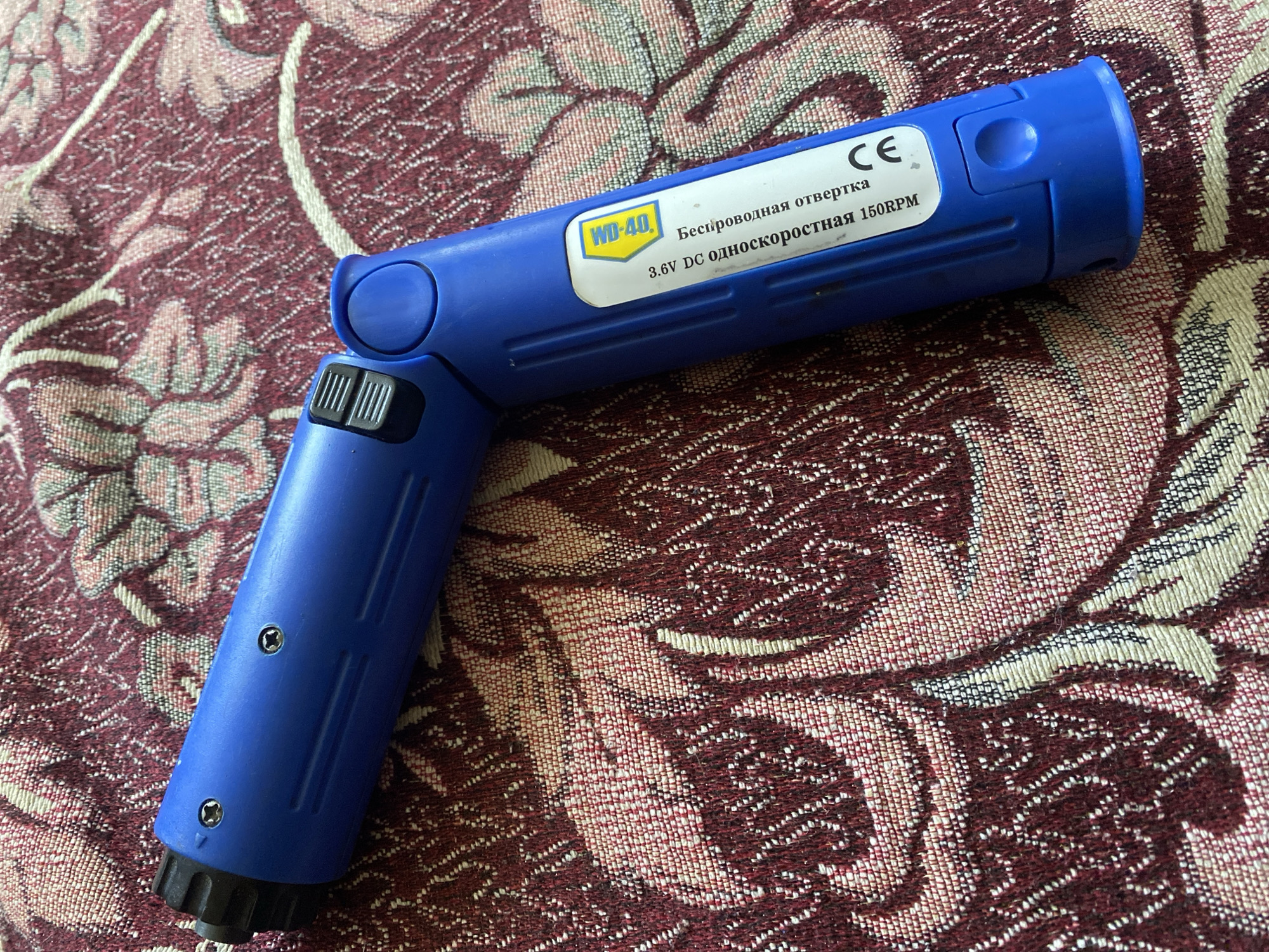 A blue cordless screwdriver is lying on a patterned fabric. The screwdriver is L-shaped and has a black textured grip at one end. A white label with a yellow "WD-40" logo is attached to the body of the screwdriver. The label has text in Russian which translates to "Cordless screwdriver 3.6V DC 150RPM" and the "CE" mark. The fabric appears to be a tablecloth or similar textile with a floral pattern in shades of maroon and cream. The image was taken in Kursk, Russia.