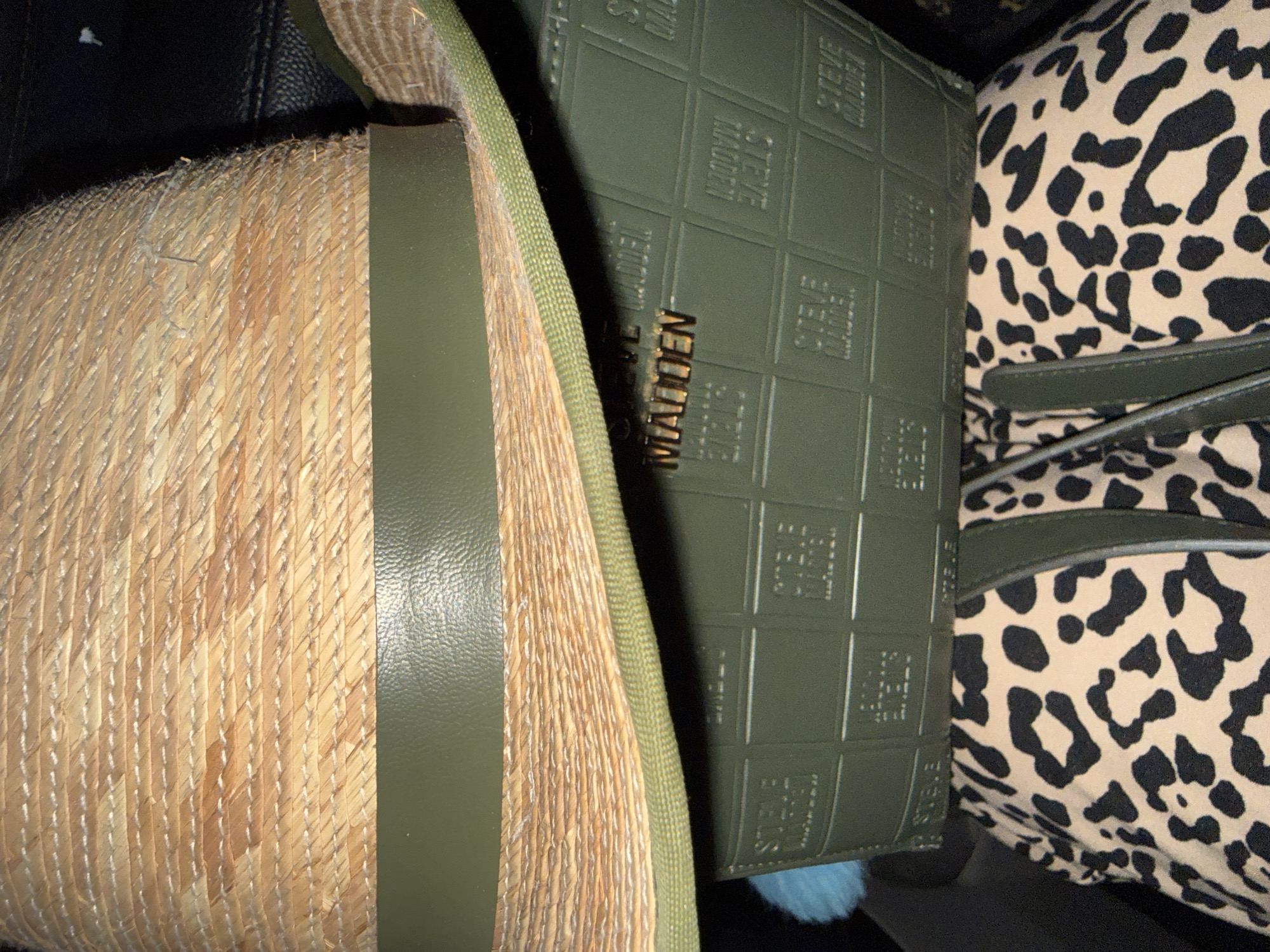 The image shows a close-up view of a straw hat with a green leather band. Below the hat is a green clutch bag with an embossed "Steve Madden" logo repeated in a grid pattern. A piece of leopard-print fabric is visible at the bottom of the image, possibly part of an outfit or another accessory. The overall scene appears to be a flat lay or a still life arrangement. The lighting suggests an indoor setting, and the background is dark and out of focus. There are no people or discernible locations in the city of Kennesaw, United States of America.