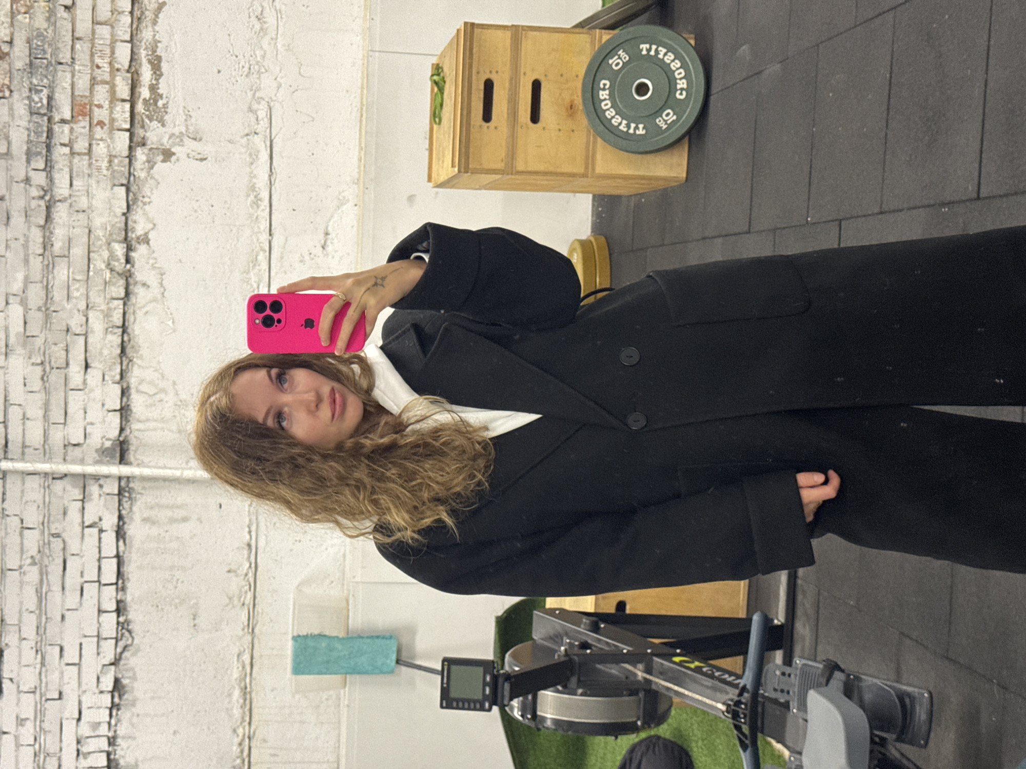 A young woman is captured taking a selfie in an indoor setting, likely a gym or functional fitness facility. She has long, wavy, light brown hair and blue eyes, looking directly at the camera with a neutral expression. She is wearing a dark, possibly black, long coat over a white top or hoodie. Her right hand, adorned with a ring and a small tattoo, holds a bright pink smartphone, likely an iPhone given the distinct camera array and visible Apple logo on the case.
The background reinforces the gym environment. To the right, a wooden plyometric box sits near a green weight plate, which displays the text "CROSSFIT" and "10 KG" (both reversed as seen in the selfie). Below the woman, parts of a Concept2 rowing machine are visible, including its monitor and flywheel. The floor is covered in dark, possibly rubber, gym mats. The walls feature a textured, industrial look, with sections of exposed, light-colored brickwork and painted concrete showing signs of wear. A small, light blue towel or mat is also visible on the floor to her left. The lighting appears to be artificial, typical of an indoor space.