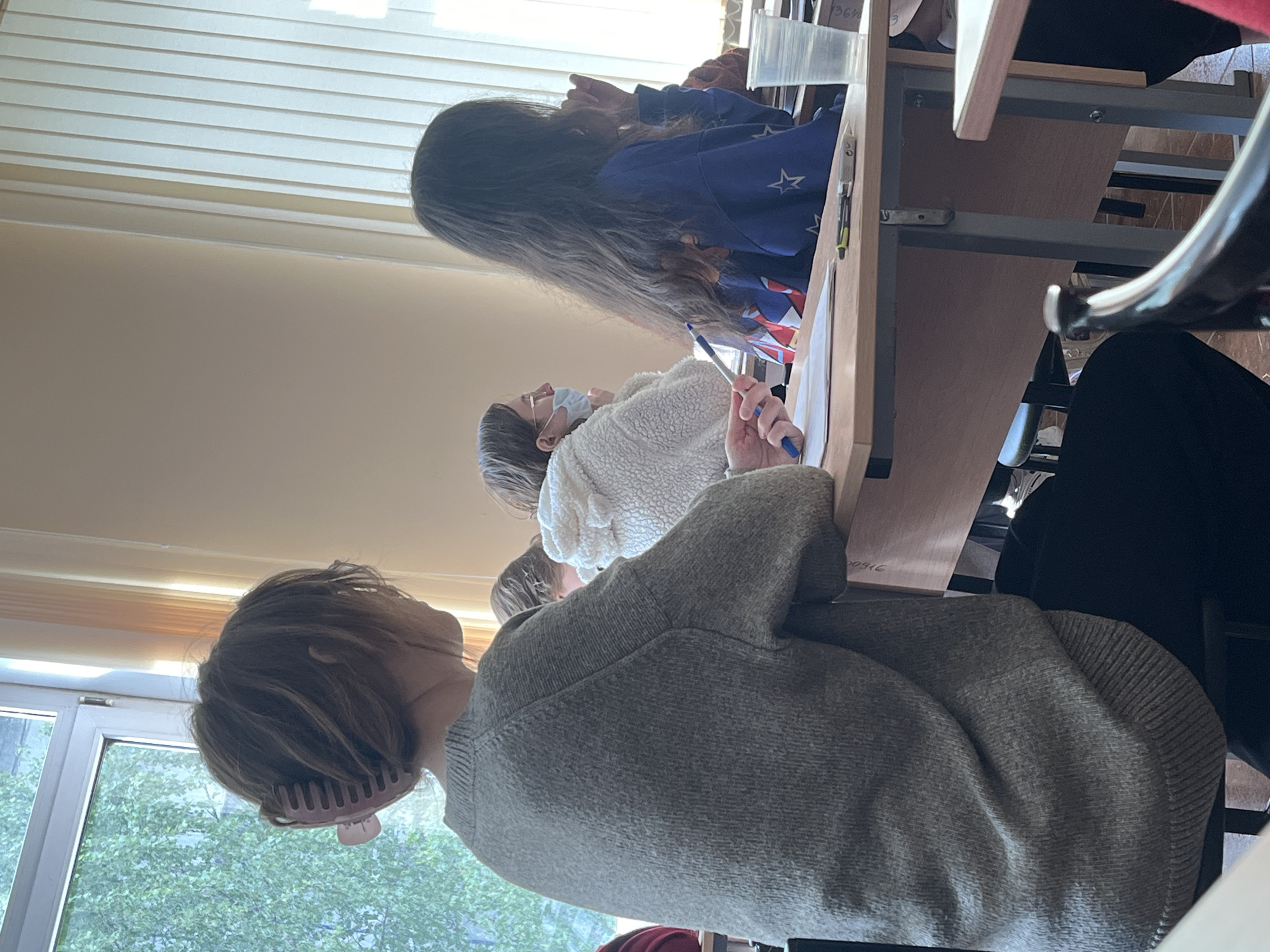 In a room, possibly in Saint Petersburg, Russia, several people are seated at desks, seemingly students in a classroom. The individual in the foreground has short hair and is wearing a grey sweater. Another person, wearing a white sweater and a face mask, is holding a pen over some papers on their desk. Further away, a third person with long hair is wearing a blue sweatshirt. The room is illuminated by natural light filtering through a window. The desks appear to be made of light wood.