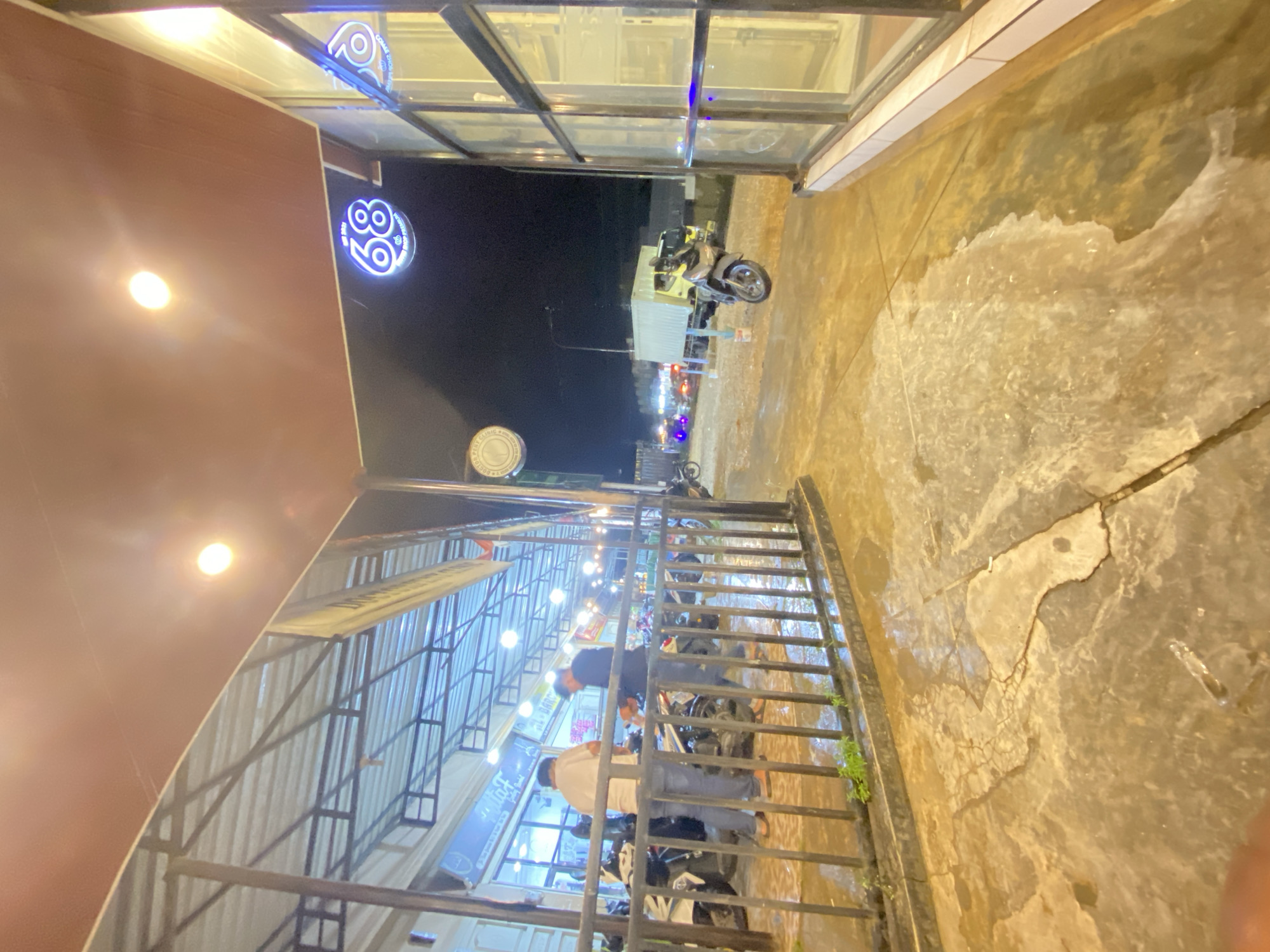 This image, presented sideways as if rotated 90 degrees clockwise, captures a vibrant night scene in Keudeublang, Indonesia, with visibly wet ground suggesting recent rain.
To the left (which would be the top if rotated vertically), a covered outdoor area features a brown ceiling with bright light fixtures illuminating the space. Above, glass panels reflect light. A prominent blue neon sign, shaped like a circle, displays the number "89" and the text "COME JOIN US". Below it, a smaller circular sign is partially visible, showing "CLINIC" and "BOBBY".
Underneath this canopy, a black metal railing separates the space from the foreground. Several motorbikes are parked in this area. At least two men are visible near the railing and motorbikes; one wears a dark shirt, the other a white shirt, both engaged in indistinct activities, possibly conversing or preparing to leave. More figures can be seen deeper within the covered area, which appears to be a cafe or gathering spot with tables and lights.
The immediate foreground and extending to the right (the bottom if rotated vertically) show a cracked concrete or paved ground, extensively wet with reflective puddles and damp patches. Small sprigs of green grass grow along the base of the metal railing. Beyond the railing, under the dark night sky, streetlights glow in the distance. A motor-scooter is parked on the wet ground, and further down the street, a light-colored box truck is visible. The numerous artificial lights scattered across the scene create a lively, urban atmosphere at night.