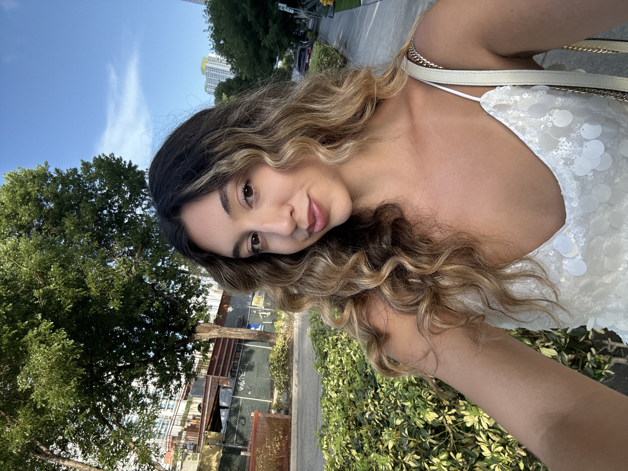 A young woman with long, wavy, highlighted brown hair is taking a selfie outdoors. She is wearing a white sequined top with spaghetti straps and a white purse with a gold chain strap slung over her right shoulder. The background shows a sidewalk, a street, lush green bushes, a large tree with green leaves, a chain-link fence, and buildings, including some high-rise apartments in the distance. The sky is blue with a few wispy clouds. The scene appears to be in Miami, Florida, USA.