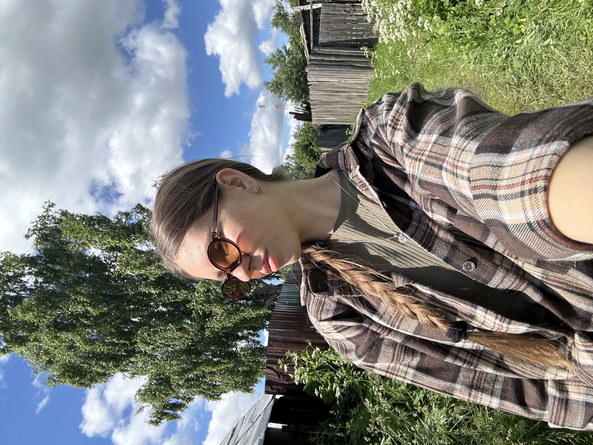 In this medium shot, a young woman with long, braided brown hair and brown-rimmed sunglasses is pictured from the chest up. She is wearing a brown and beige plaid shirt over a green ribbed t-shirt and a silver heart pendant necklace. The woman is outdoors in what appears to be a rural setting. A large green tree stands to her left, and a wooden fence stretches across the background. The sky is blue with scattered white clouds. The overall scene suggests a sunny day. The location is in the village of Malaya Borshchevka, Russia.