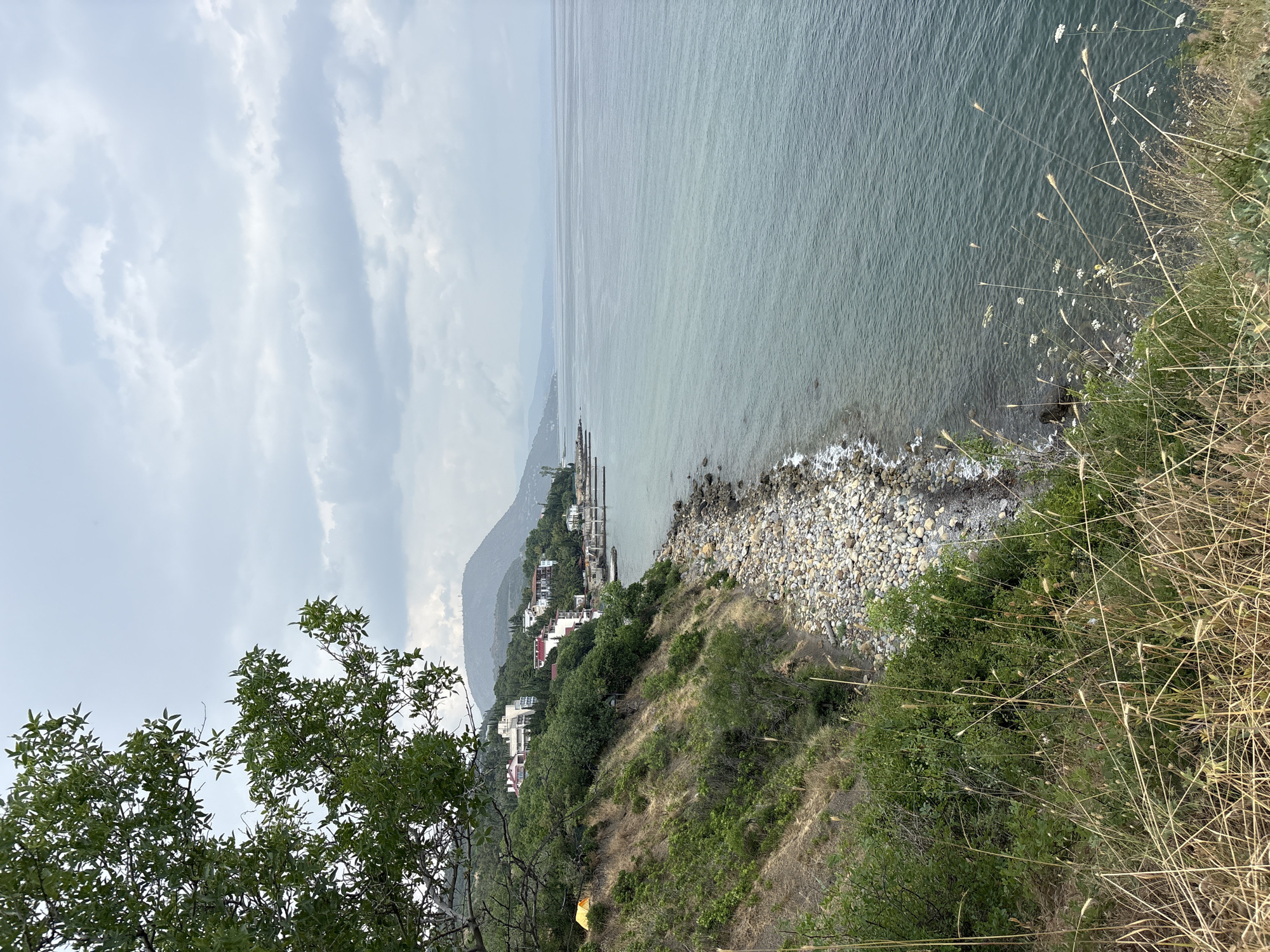 The image depicts a coastal scene with a slightly overcast sky. In the foreground, a dense growth of dry grass and some green foliage is visible. A steep, vegetated slope descends towards a pebble beach. Along the crest of the slope, several buildings, appearing to be a resort or residential complex, are situated among trees and greenery. The sea stretches out to the horizon, with gentle waves lapping the shore. A thin line of distant mountains can be seen along the coastline. A small, bright yellow tent is pitched on the slope, near the edge of the vegetation. The scene is devoid of people. The location is identified as Bondarenkovo, Ukraine.