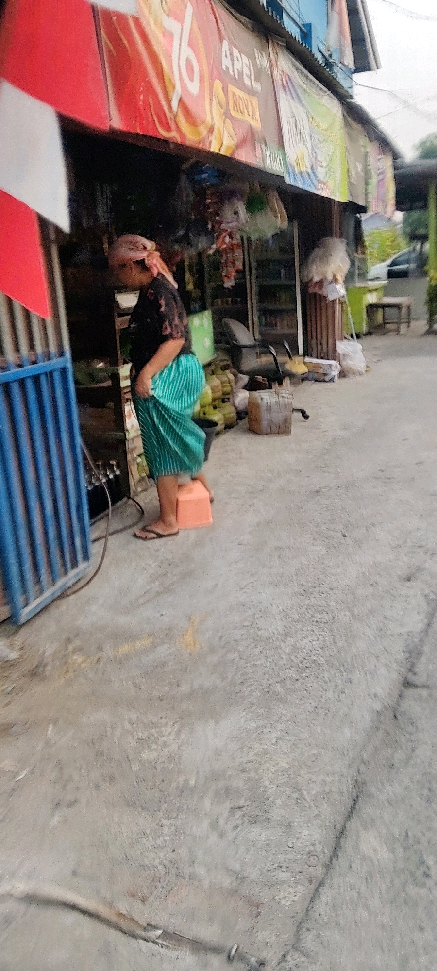 The image depicts a street scene in Mekarmukti, Indonesia, featuring a woman standing on a small orange stool in front of a shop. She is wearing a dark patterned shirt and a green striped skirt, with a headscarf. The shop's facade is adorned with red and white banners, one featuring a prominent "76" and text that appears to read "APEL ROVA". Shelves inside the shop are stocked with various items, including what appear to be bottled drinks and other products. To the left of the woman, a blue metal gate is visible. The ground is paved with concrete, showing some wear and tear. In the background, there are more shopfronts, a parked car, and a table with chairs. The overall impression is of a commercial street in a local Indonesian town.