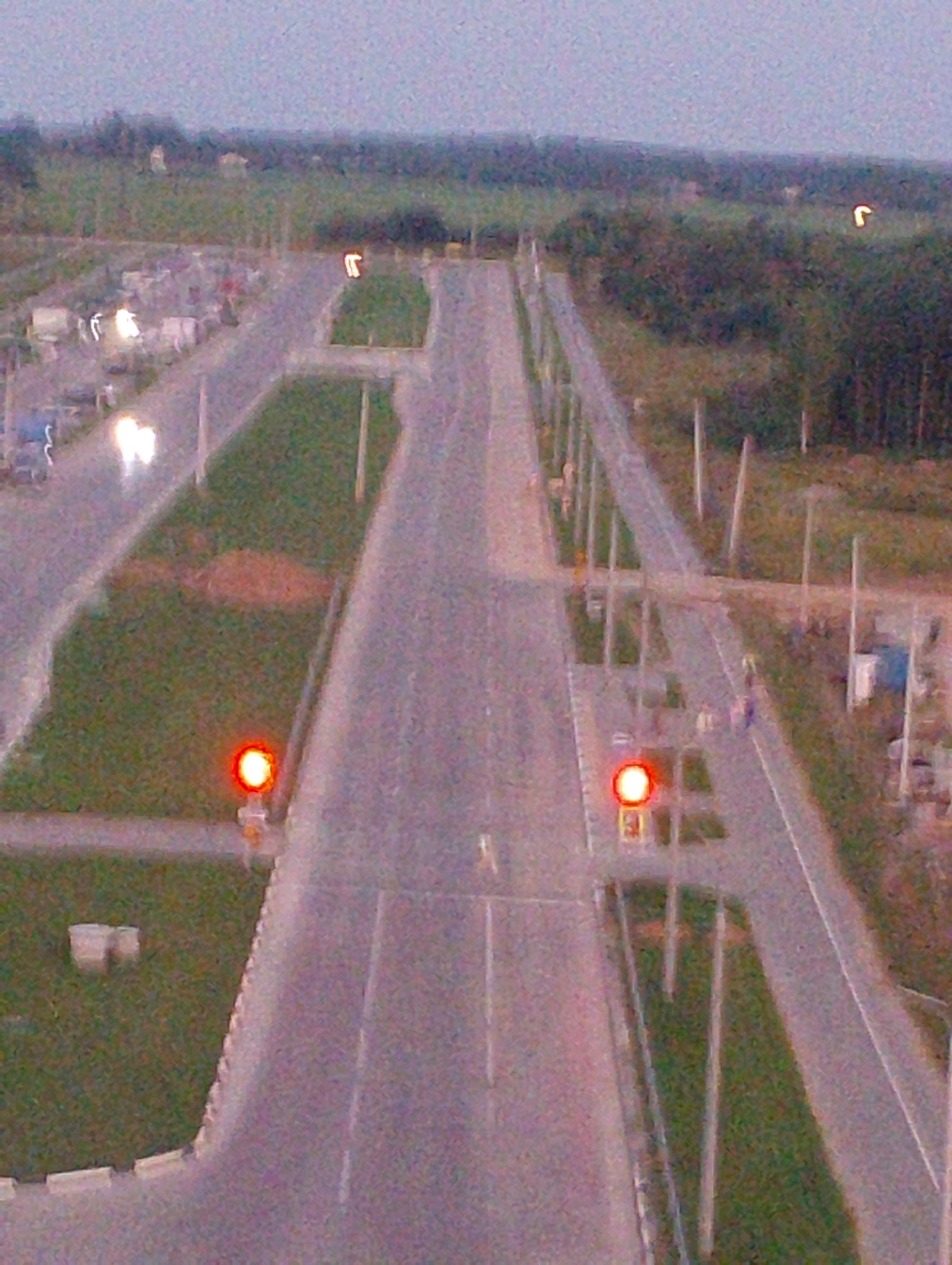 An aerial, dusk shot shows a grey asphalt road with white lane markings, leading through a semi-rural landscape. The road is flanked by grassy verges and occasional trees. There are also street lights and traffic signals visible. The image is grainy and somewhat blurry, but appears to show a scene in Vitsebsk, Belarus. A car with its headlights on is visible in the distance, and there is a building in the background.