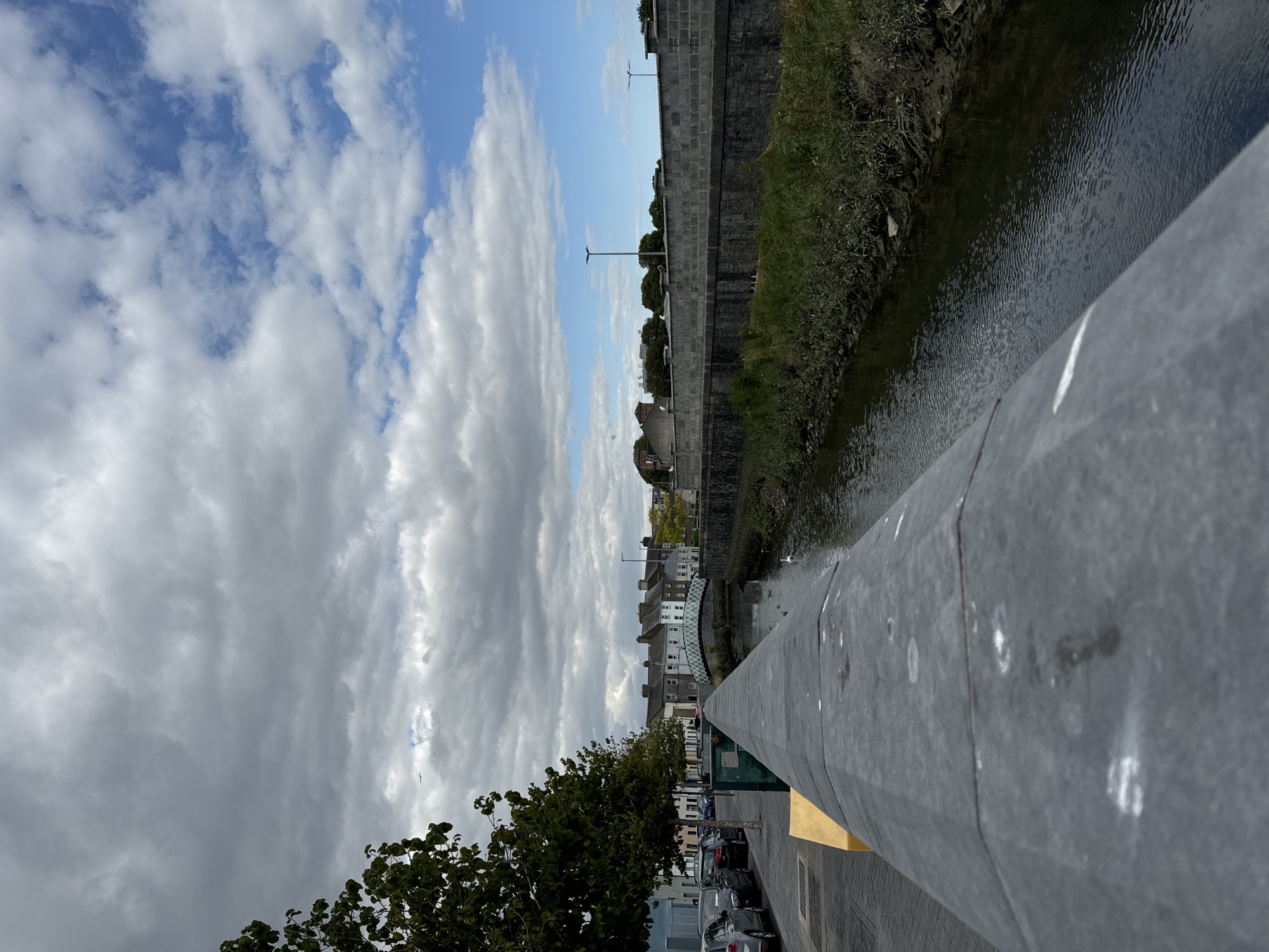 A view of the River Suir in Waterford City, Ireland, looking towards the west. The foreground is dominated by a dark grey stone parapet that occupies the lower left portion of the image, extending into the distance. Beyond the parapet, a sidewalk with parked cars is visible to the left, and a narrow canal or river flows to the right, flanked by grassy banks. Across the river, a row of terraced houses with white-trimmed windows line the opposite bank, and a decorative white bridge spans the water further downriver. The sky above is a mix of dramatic grey clouds and patches of bright blue.