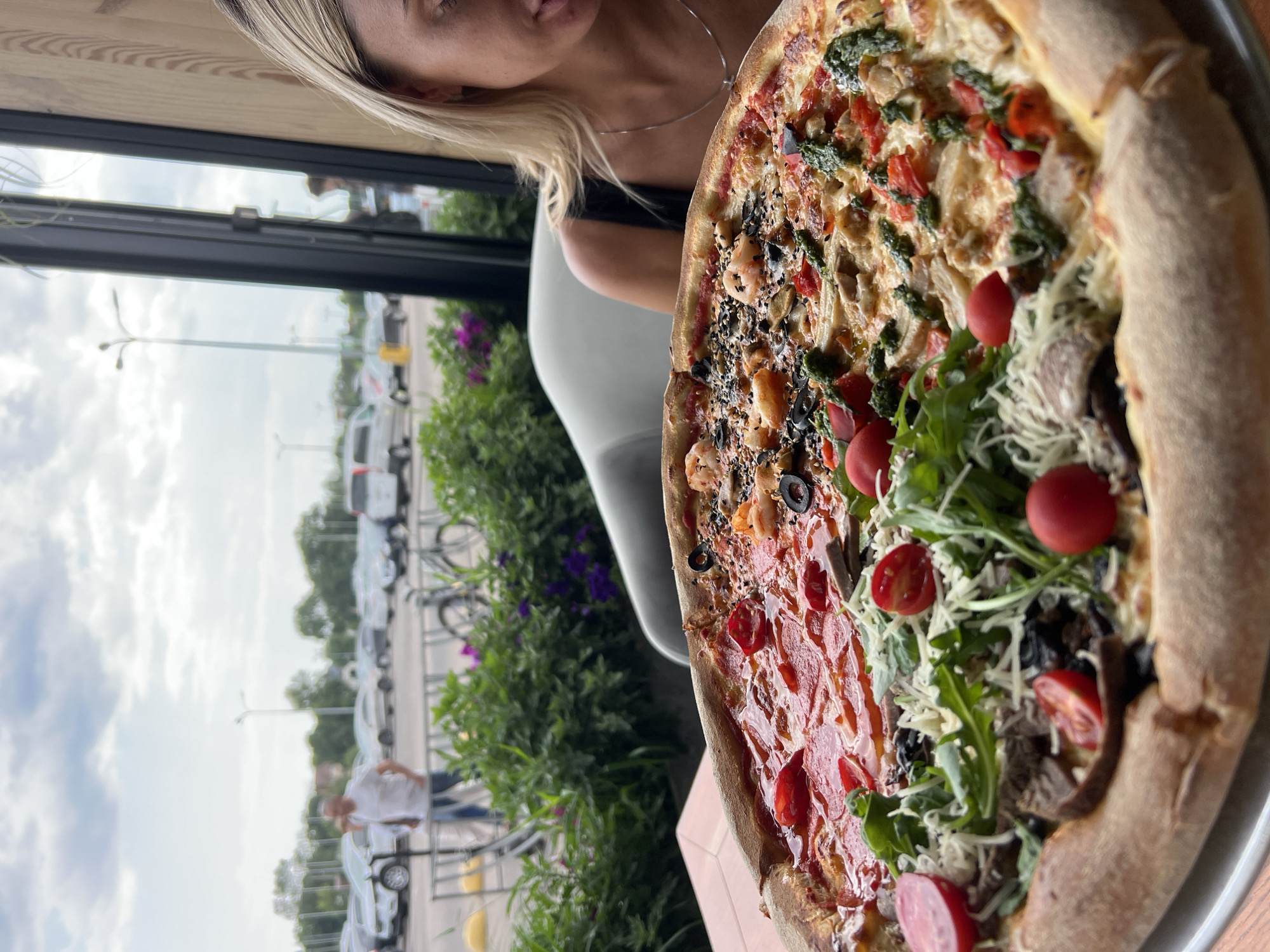 A large, multi-topped pizza is presented in the foreground, positioned on a circular metal tray. The pizza is divided into distinct sections, each featuring different toppings. One section displays pepperoni and cherry tomatoes. Another section is topped with shrimp, olives, and what appears to be sesame seeds. A third section includes mushrooms, arugula, shredded cheese, and cherry tomatoes, with visible pieces of meat. The final section of the pizza is topped with chicken or other white meat, tomatoes, and green sauce.
In the background, beyond a window, a parking lot is visible, with several cars and a light pole. A man in a white t-shirt and blue jeans is walking in the parking lot, and a bicycle is parked near some green foliage.
To the right of the pizza, a woman with blonde hair and a black top is partially visible. She is seated and looking down, seemingly towards the pizza. The setting appears to be an outdoor or semi-outdoor dining area, possibly a restaurant with a view of the parking lot and some greenery. The overall impression is a casual dining scene featuring a substantial and varied pizza. The location is stated to be Fontanka, Ukraine.