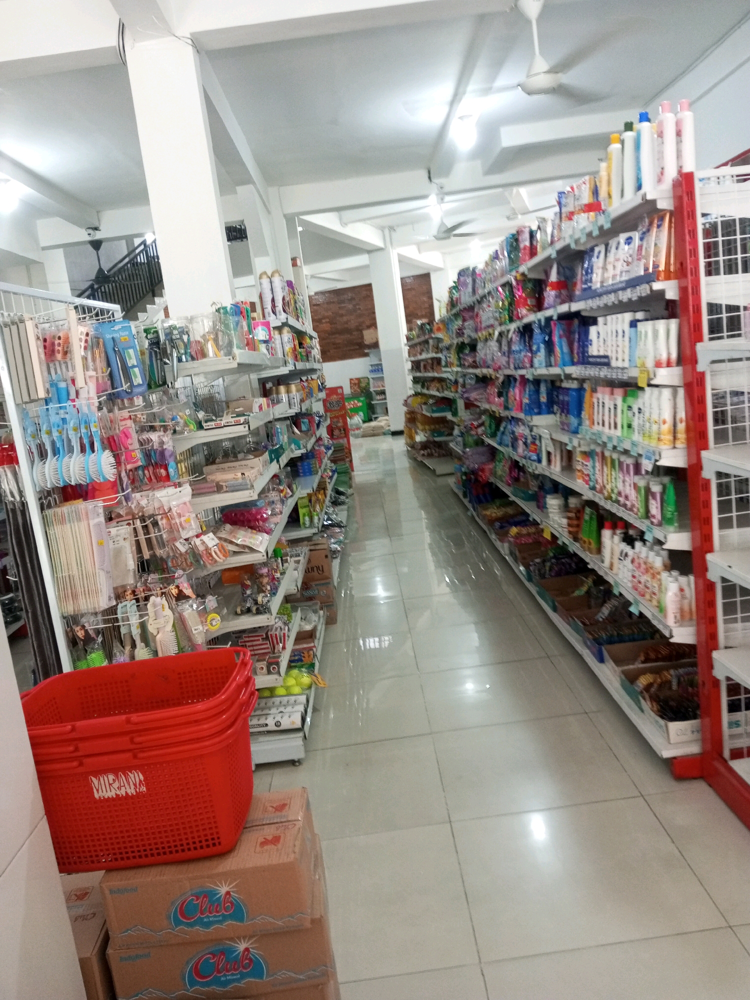 The image depicts the interior of a retail store, likely a minimarket or supermarket, in Malang, Indonesia. The store features aisles stocked with various consumer goods on shelves. On the left side of the image, shelves are filled with household items, cleaning supplies, personal care products, and toiletries. A prominent red shopping basket is visible in the foreground, placed on top of stacked cardboard boxes. The floor is tiled and appears clean and well-maintained. On the right side, shelves are lined with a wide array of personal care products, including shampoo bottles, soaps, and lotions, as well as other packaged goods. The store is brightly lit, with overhead fluorescent lights and ceiling fans providing illumination and air circulation. The overall environment suggests a typical commercial establishment designed for everyday shopping.