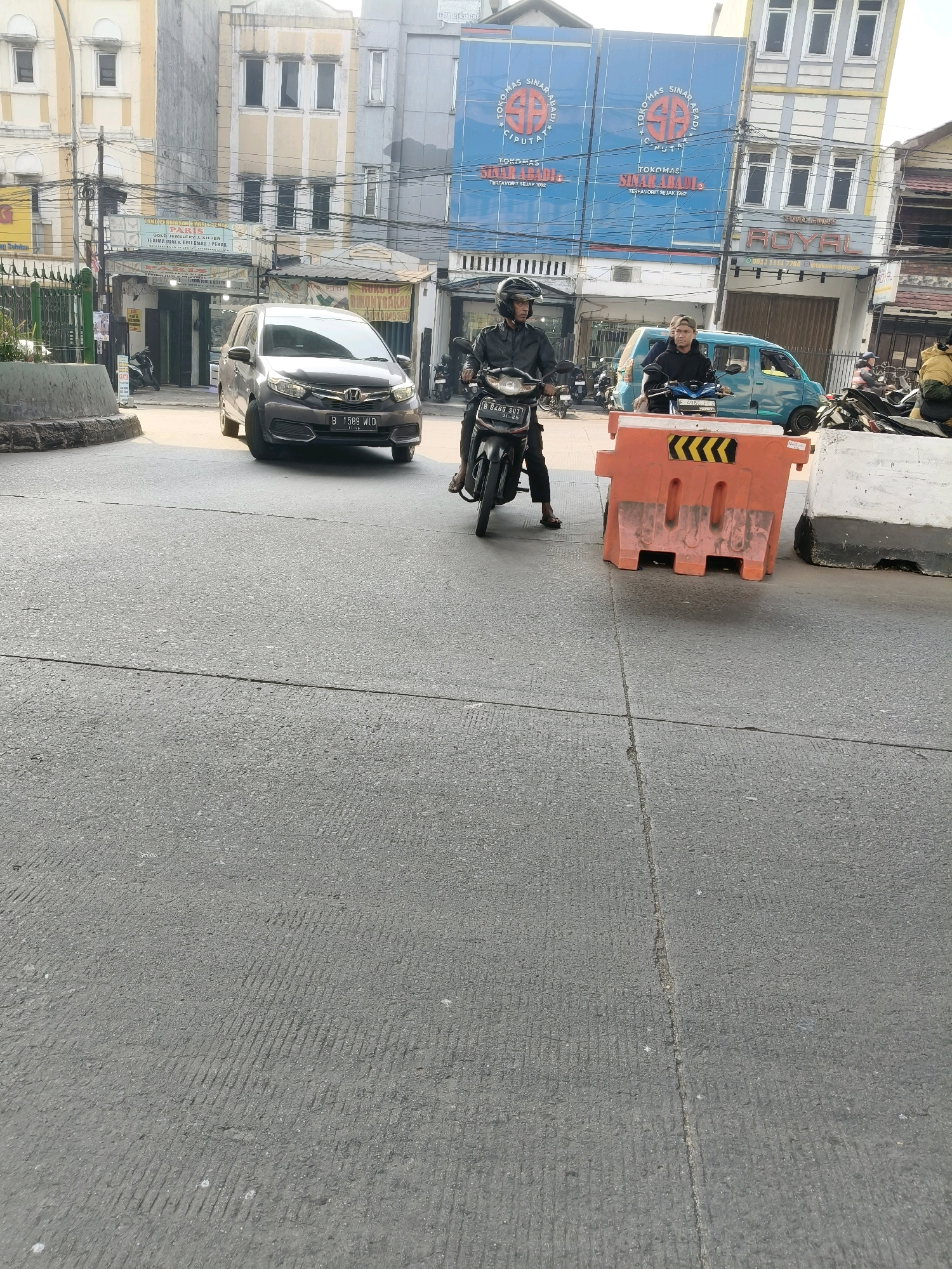 The image shows a street scene in South Tangerang, Indonesia. In the foreground, a grey car with license plate B 1599 WID is stopped at a traffic light. The car is facing right. To the right of the car is an orange traffic barrier. Behind the car, two men on motorcycles are stopped, also at the traffic light. The man on the left is wearing a black jacket and helmet, and the man on the right is wearing a black hoodie and a cap. The background shows several buildings, including a blue building with text that appears to be store signage. The street is paved with asphalt. The weather appears to be clear and sunny.