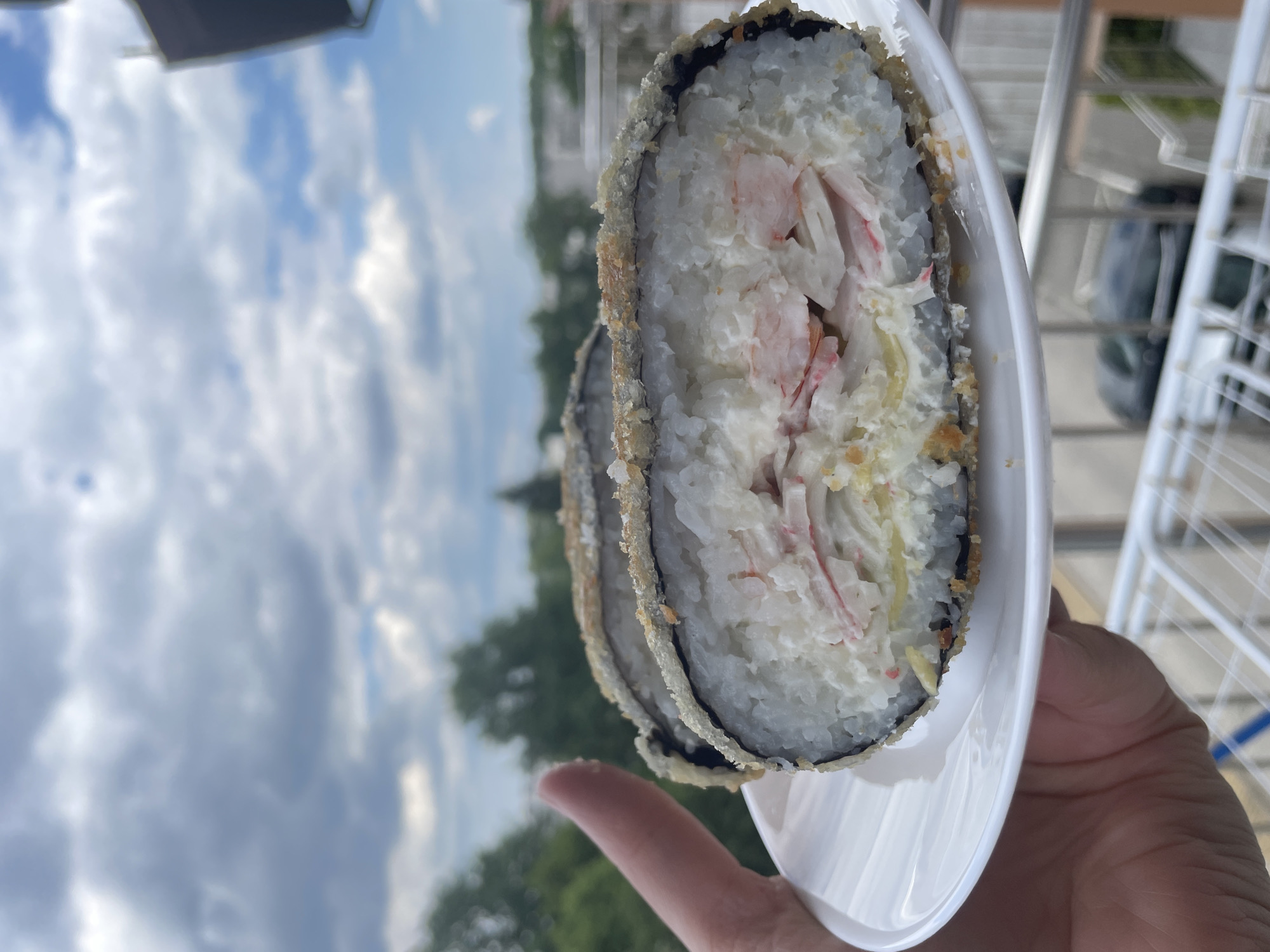 A close-up, over-the-shoulder shot shows a person's hand holding a white plate with two pieces of fried sushi rolls. The sushi appears to be tempura fried, with a crispy outer layer and white rice. The filling is visible, consisting of what looks like imitation crab meat, cream cheese, and possibly avocado or cucumber. The background is blurred, showing a cloudy sky with patches of blue and green trees in the distance, suggesting an outdoor setting in a city. The person's thumb is visible on the left side of the frame, partially obscuring the plate. The lighting suggests it is daytime, with soft, diffused light.