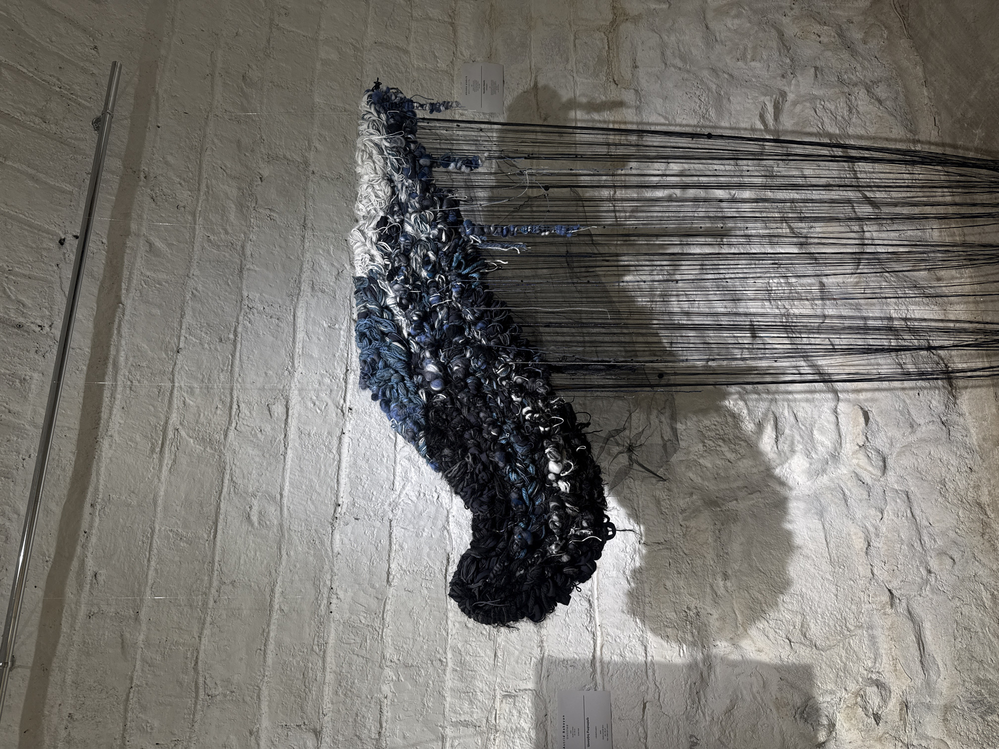 The image displays an indoor textile art installation within an exhibition space. The primary subject is a large, organic-shaped sculpture composed of densely packed yarns and fabric scraps, approximately 1.5 meters wide and 1 meter high, suspended against a textured wall. The sculpture exhibits a color gradient, transitioning from dark navy blue and black on its left side to lighter blues, grays, and white towards its right. Numerous thin, dark, vertically hanging threads descend from the underside of the main textile mass, extending downwards beyond the frame, with a few thicker, shorter, blue-toned strands interspersed among them.
The artwork is suspended by thin, transparent monofilament lines from a silver-colored metal track secured to the ceiling. The background wall is constructed from rough, irregular stone blocks, painted an off-white color, revealing significant texture and mortar lines. The upper section of the wall curves into an arched ceiling. Overhead illumination casts pronounced shadows of the artwork and its suspension track onto the textured wall, accentuating its relief.
Two white rectangular information plaques are affixed to the wall. The plaque on the left identifies the artist as "Astrid Bekayan" and the artwork as "Untitled," with the same information provided in Armenian (Աստրիդ Բեգոյան / Անվերնագիր). A second, partially visible plaque on the right also names "Astrid Bekayan" and "Unbroken Silence." Based on the artist's context, the exhibition space is located within the city of Yerevan, Armenia. No individuals are present in the scene.