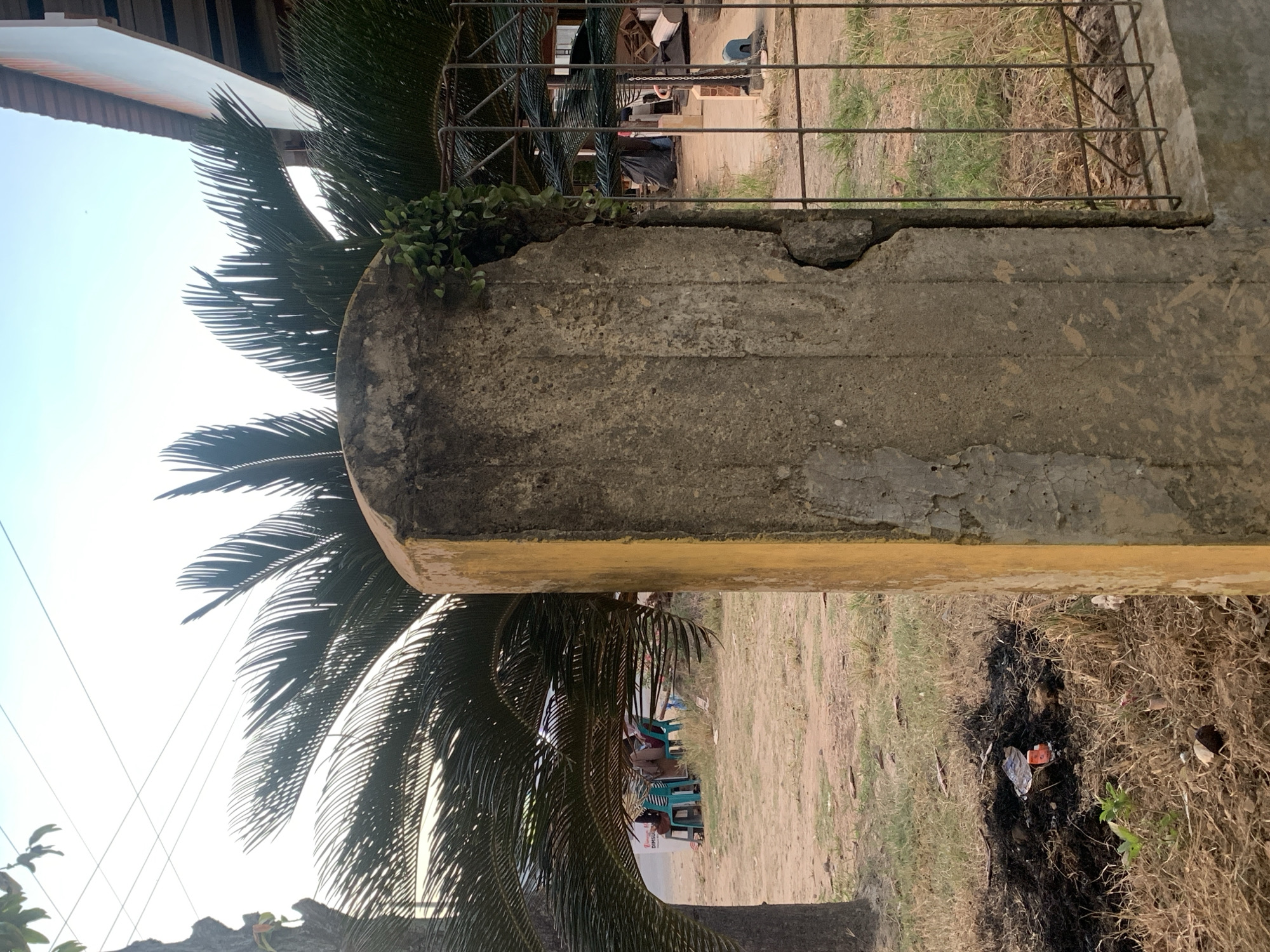 This is a photograph taken in Kualadua, Indonesia, at ground level, depicting a concrete post in the foreground. The post is partially covered by yellow paint and shows signs of damage, with a large crack running down its right side. Green vines are growing out of this crack. Behind the post, a palm tree is visible, its fronds casting shadows. In the background, there are several plastic chairs set up, with people sitting on them, suggesting a gathering or a place of rest. A road is visible beyond the chairs, and further in the distance, a building with a roof is present. The ground appears to be dry and dusty, with some patches of grass and evidence of burnt material in the lower left corner of the image. A metal fence runs along the right side of the concrete post. The sky is clear and bright.