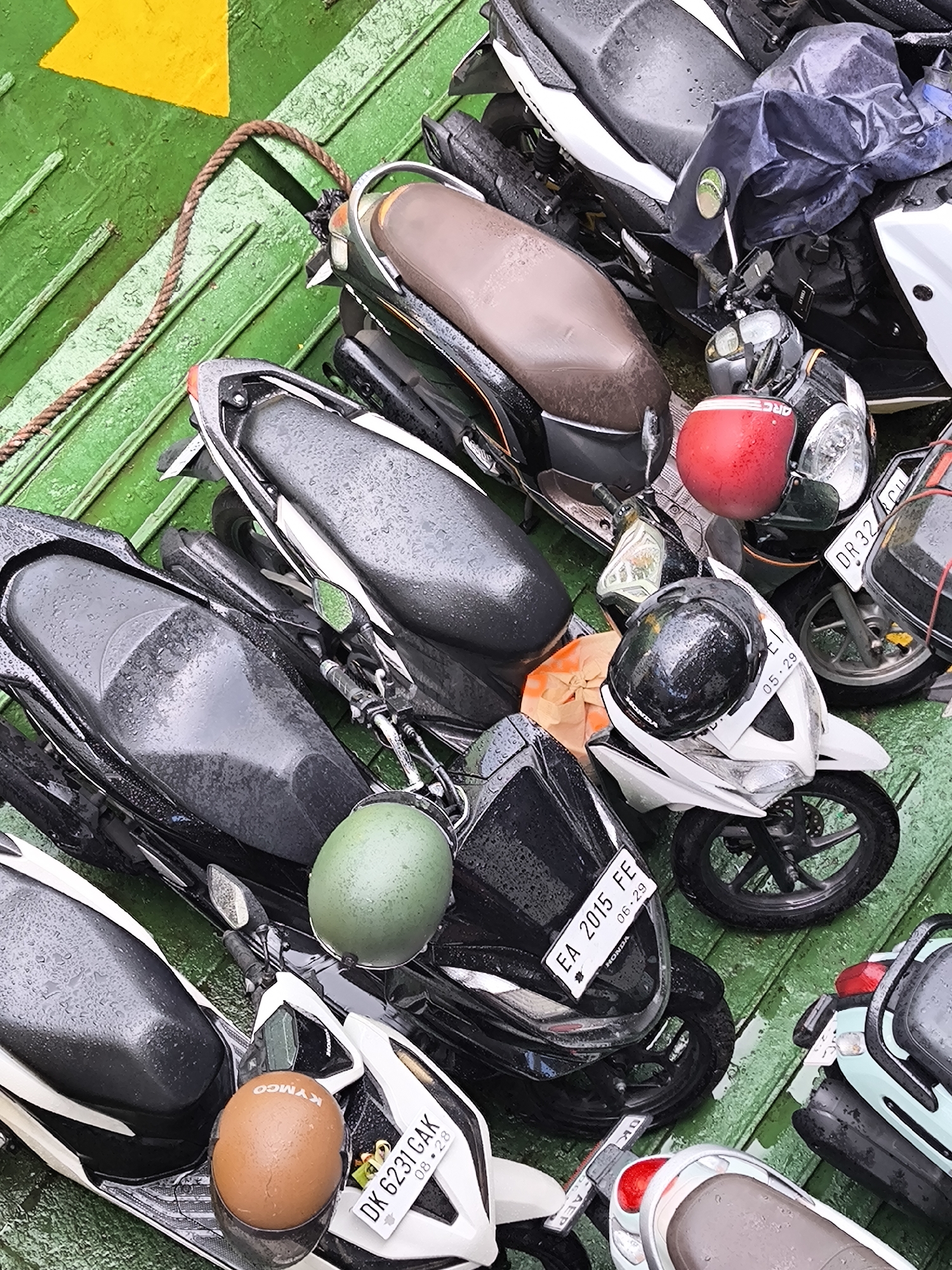 An overhead, high-angle shot captures a crowded scene of numerous scooters and helmets tightly packed together on a wet, green, ribbed surface, likely the deck of a ferry. The green floor features a yellow arrow marking and a thick brown rope running along one side. Water droplets are visible on all surfaces, indicating recent rain or spray.
The scooters are a mix of models and colors, predominantly black and white, with one light blue/green scooter visible at the bottom right. Several helmets are resting on the scooters: a bright red one on a black scooter, a glossy black one on a white scooter, a matte green one on another black scooter, and a brown/orange one with "KYMCO" branding visible on a white scooter. "HONDA" logos are discernible on some of the white scooters.
Visible Indonesian license plates provide location cues: "EA 2015 FE" with "06·29" below it is on a black scooter; "DK 6231 GAK" with "08·28" is on a white scooter; a partial "DR 32.CUI" is seen on a scooter at the top right; and another plate shows "05·29". The tight packing suggests the scooters are being transported, likely on a ferry between islands in Indonesia. The scene depicts a common mode of transport for vehicles in the region.