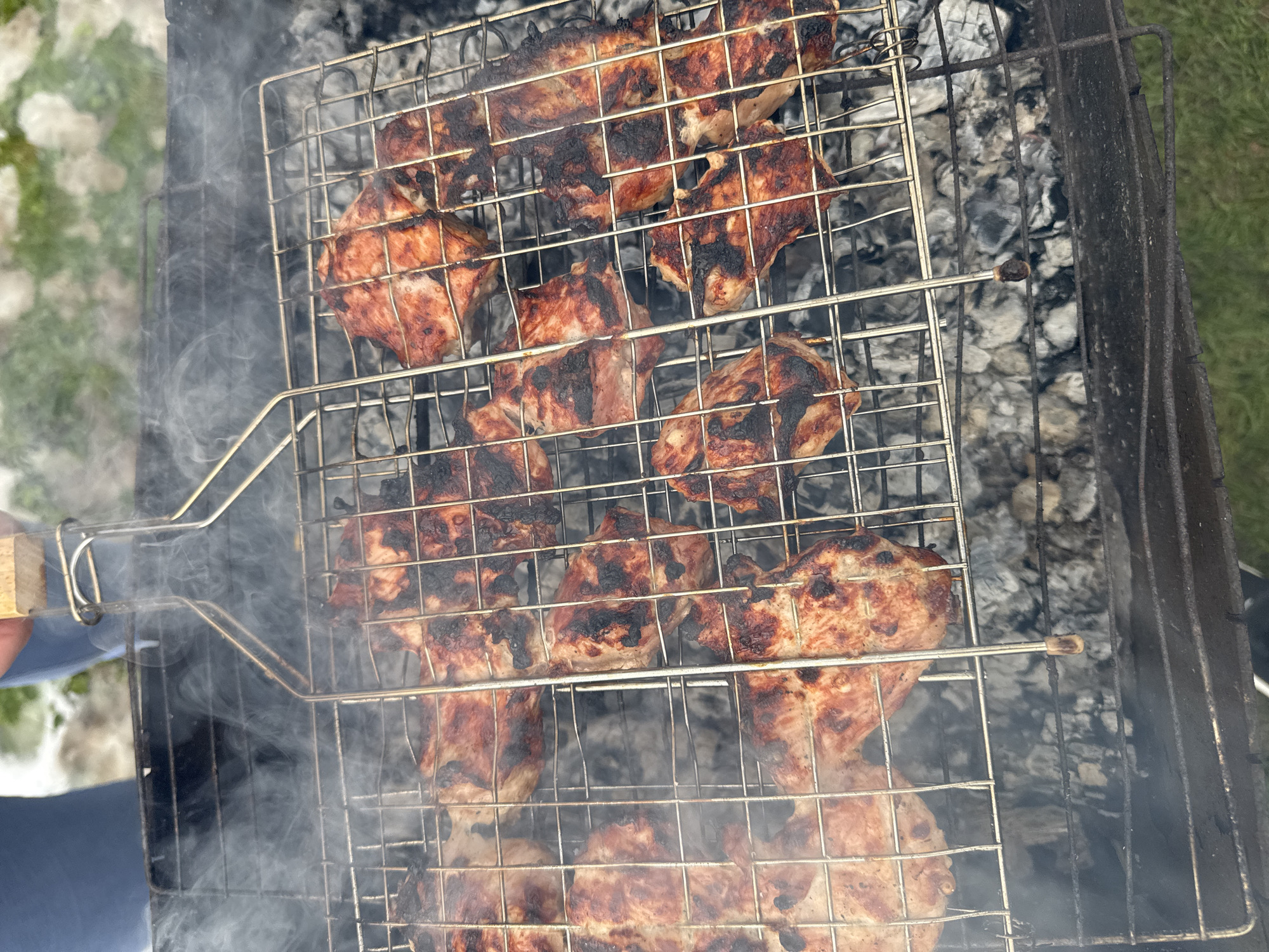 This image depicts an outdoor barbecue scene in Раздолье, Russia, with a focus on meat being grilled over charcoal. The central subject is a metal grilling basket, filled with numerous pieces of seasoned meat, likely chicken or pork, which are visibly cooking. The meat displays charring and grill marks, indicating it is well into the cooking process. Thick white smoke billows upwards from the hot coals beneath the grill, partially obscuring the background and adding to the atmosphere of active cooking.
The meat is held within a hinged metal grill basket, with a person's hand, partially visible in the lower-left, holding the wooden handle. The grilling basket is positioned over a dark, rectangular charcoal grill, or mangal-style grill, filled with grey ash and glowing embers. In the background, beyond the smoke, patches of green grass are visible, interspersed with lighter areas that could be snow or dry vegetation, suggesting an outdoor setting during daytime in potentially cooler weather. The overall scene captures a casual moment of food preparation outdoors. No visible text is present in the image.
