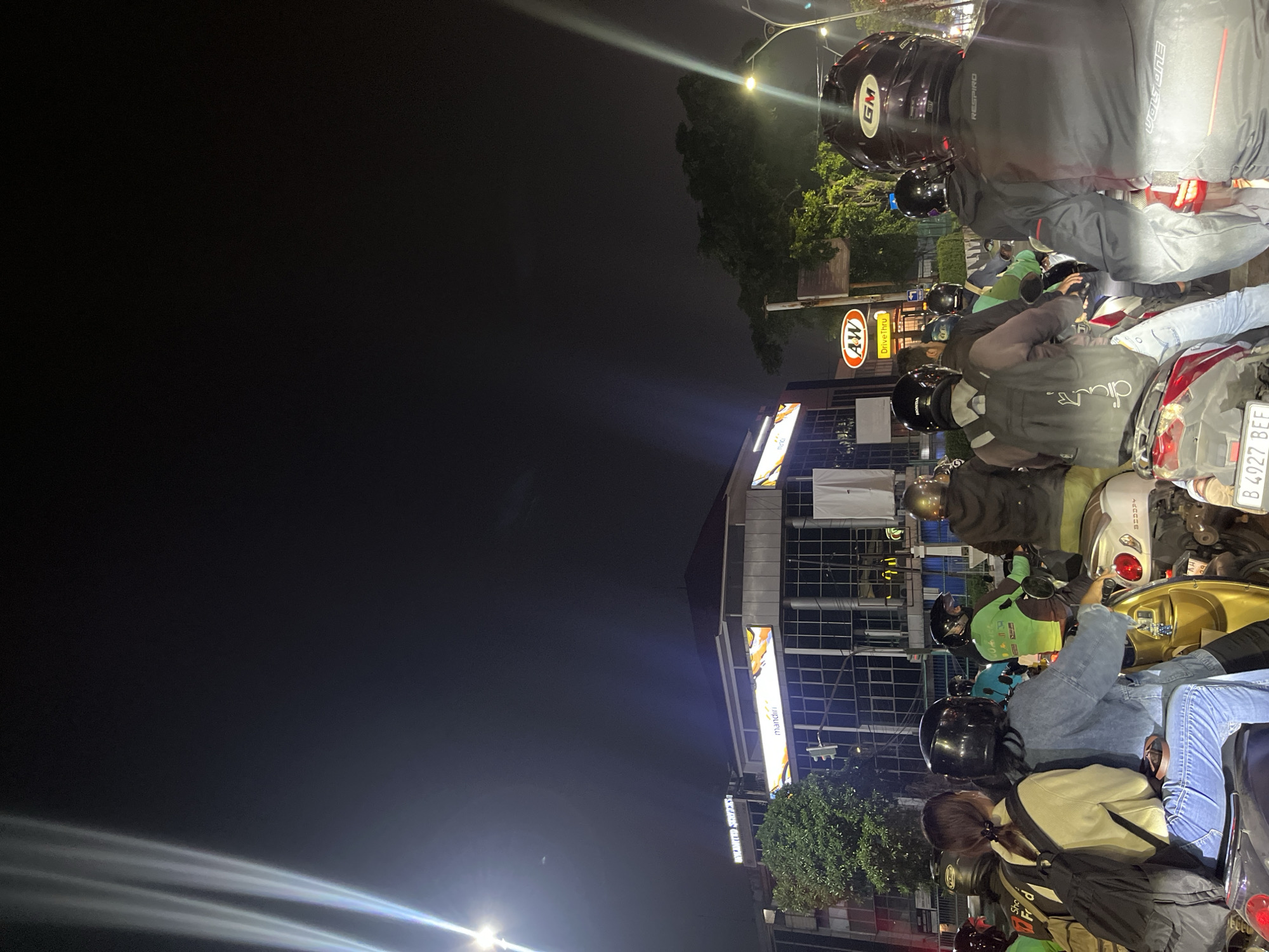 The image depicts a nighttime street scene in West Jakarta, Indonesia, likely during a traffic jam. A group of motorcyclists, predominantly wearing helmets, are stopped on the road. The foreground is dominated by the backs of several individuals on motorcycles, with one rider in a black helmet and jacket with "RESPiRO" and "VOLSONE" visible. To their left, a person in a light-colored hoodie and blue jeans is seated on a motorcycle. Further back, other riders are visible, including one in a green jacket with the "Gojek" logo and another in a black helmet.
Behind the motorcyclists, a commercial building is illuminated. A prominent sign for "mandiri" is visible on the building's facade. To the right of the "mandiri" sign, another illuminated sign displays "AW DriveThru" for A&W Restaurants. Streetlights and building lights illuminate the scene, contrasting with the dark night sky. Trees and other greenery are also present, suggesting an urban environment with some landscaping. The overall impression is a typical urban traffic scene at night in a populated area of West Jakarta.