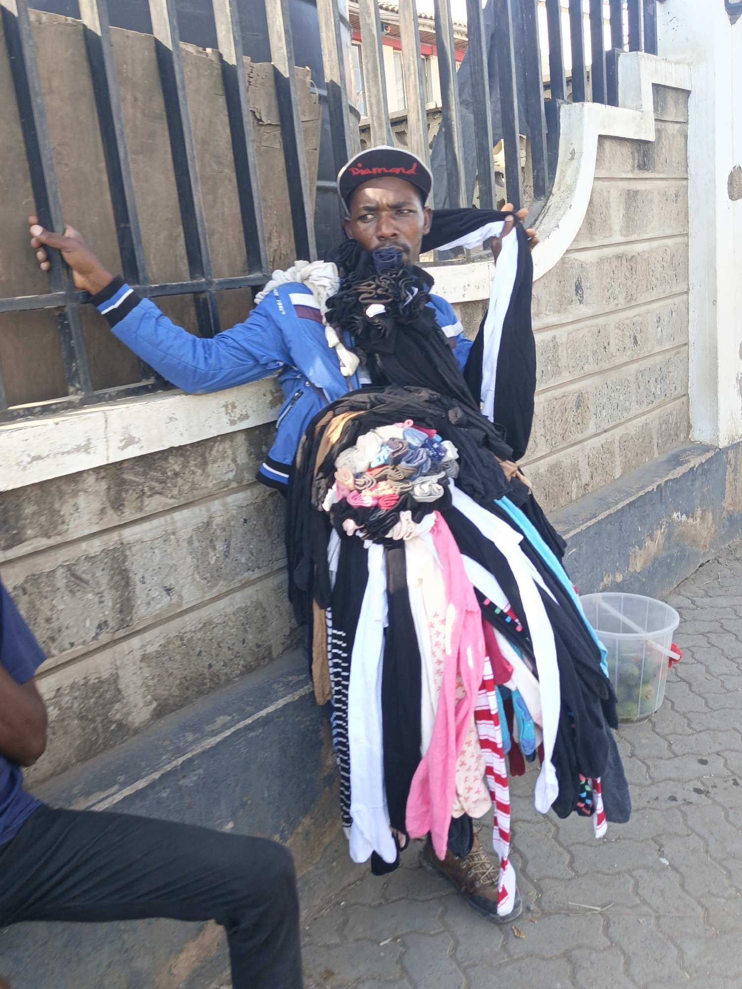 A man stands next to a wall and a black metal fence in Nairobi, Kenya. He is wearing a blue jacket, a black hat with white lettering, and brown shoes. He is holding a large pile of clothing, including black, white, and pink garments. A person is seated to the left, wearing a blue shirt and black pants. A clear plastic bucket sits on the ground next to the man. The ground is paved with interlocking stone blocks, and the wall behind him is constructed from concrete blocks.