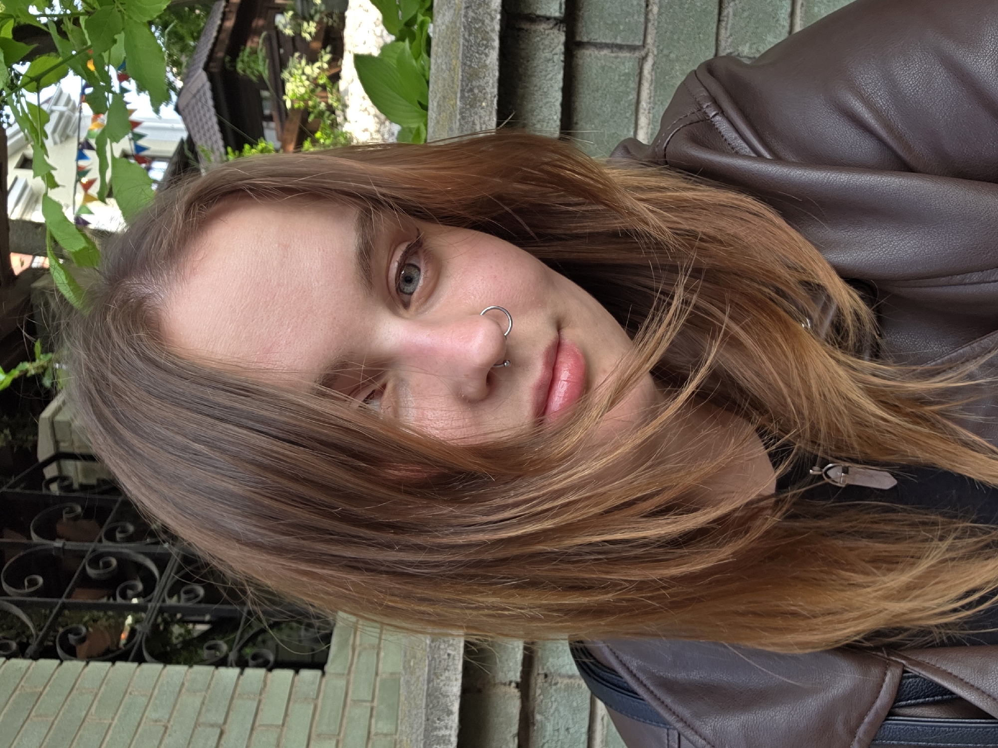 The image features a fair-skinned individual with light brown hair and a nose ring, wearing a brown leather jacket. The person is positioned outdoors, possibly in an urban setting, evidenced by the brick wall, wrought iron fence, and greenery in the background. The location is identified as the city of Unknown, Unknown.