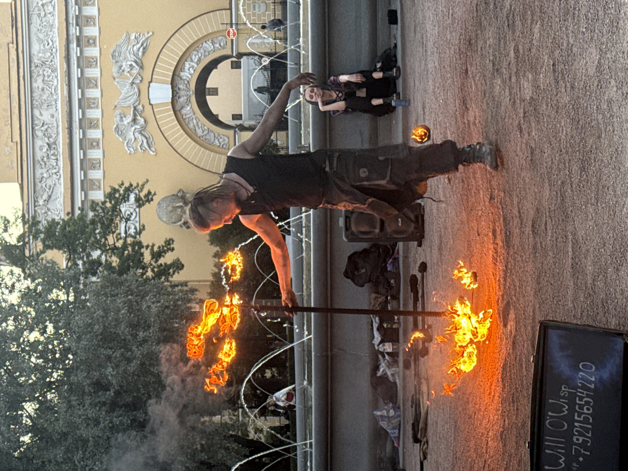 In Saint Petersburg, Russia, at an outdoor location with a fountain in the background and a building adorned with classical sculptures, a fire performer with dreadlocks is captured mid-performance. He is dressed in dark clothing and is holding a flaming prop that emits smoke. The flames are bright orange and yellow, creating a dramatic effect. In the foreground, there is a black sign with white lettering that reads "Will O'Wisp" and a phone number. To the right of the performer, a woman is sitting on a low wall, observing the show, holding a cigarette. Several other objects, including bags and what appear to be additional fire props, are scattered on the ground around the performer, suggesting a public performance space or rehearsal area. The ground is covered in gravel.