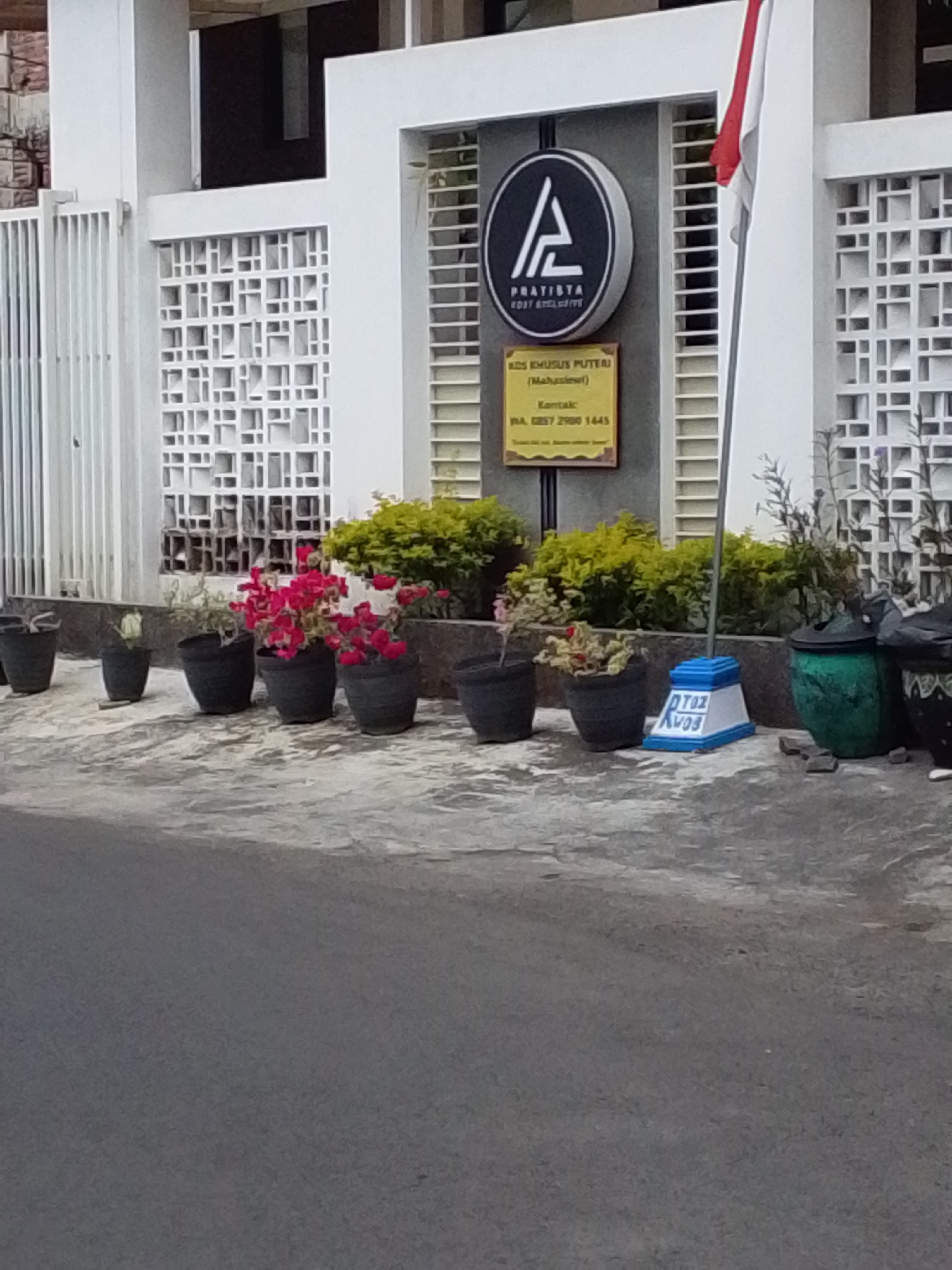 A slightly blurry, medium-shot, eye-level, outdoor photograph captures the facade of a building with a white wall and patterned window openings. A dark asphalt road is visible in the foreground, leading up to a paved area with several potted plants. In the center of the image, a circular black sign with a white letter "A" is mounted on the wall, below which is a yellow rectangular sign with text and a phone number. A red and white Indonesian flag on a pole is positioned to the right of the sign. The plants are in dark-colored pots, with some featuring vibrant pink flowers. To the far right, two large green and black pots are visible. The overall scene appears to be in an urban setting, likely in Malang, Indonesia, given the presence of the Indonesian flag and the style of architecture. There are no people visible in the image.
