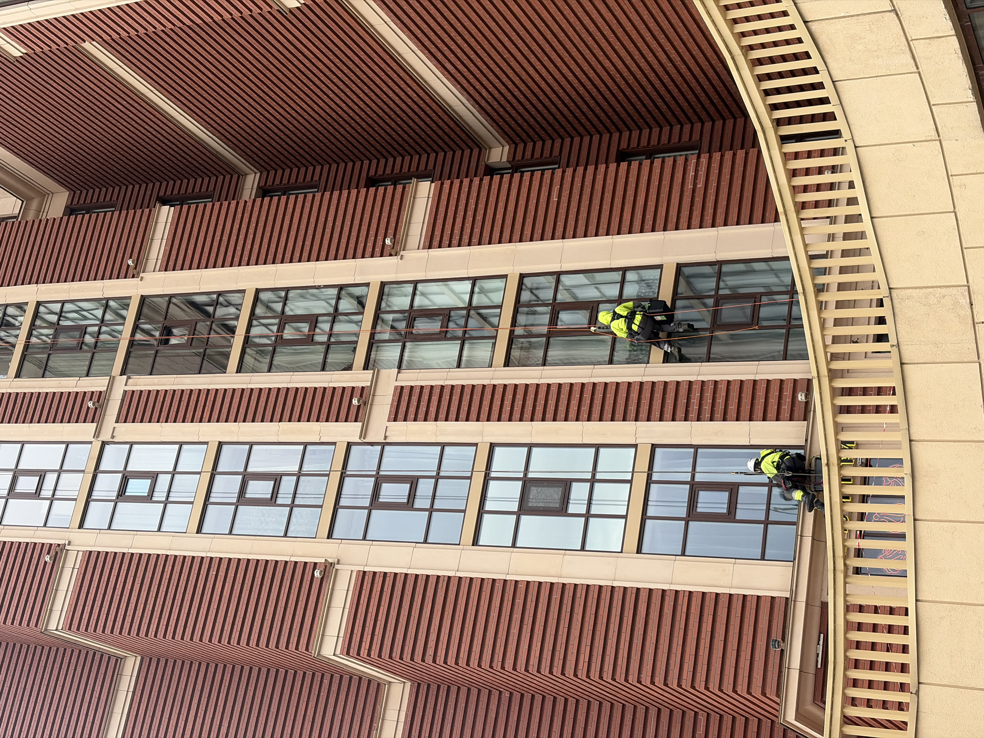 The image depicts two window washers cleaning the facade of a building in Saint Petersburg, Russia. The building has a distinctive architectural style with red vertical cladding and rows of windows. The window washers are wearing safety harnesses and reflective vests. One is seen near a curved yellow ladder.