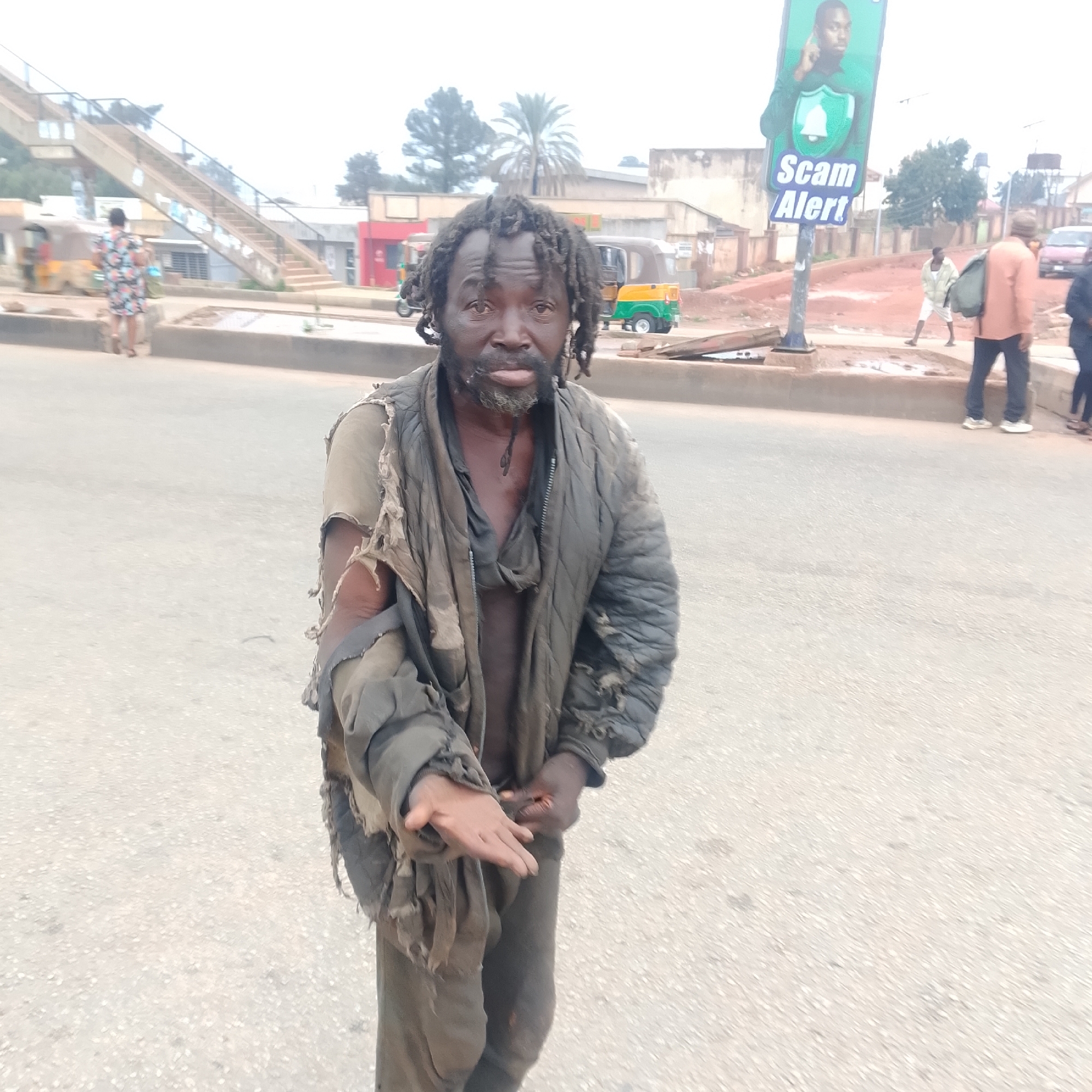 The image depicts a medium-full shot of a man with dreadlocks and a beard standing on a paved road. He is wearing tattered clothing, including a grey, quilted jacket with ripped sleeves and a dark, torn shirt underneath. His right hand is extended, palm up, as if asking for something. The man's expression is somber.
In the background, there are several vehicles, including a three-wheeled auto-rickshaw. A pedestrian overpass is visible in the upper left corner, with a person in a floral dress walking on it. A signpost with a "Scam Alert" message and an image of a person is also visible on the right side of the image. The environment appears to be an urban setting with buildings and trees in the distance. The road surface is asphalt. The lighting suggests it is daytime, possibly overcast.