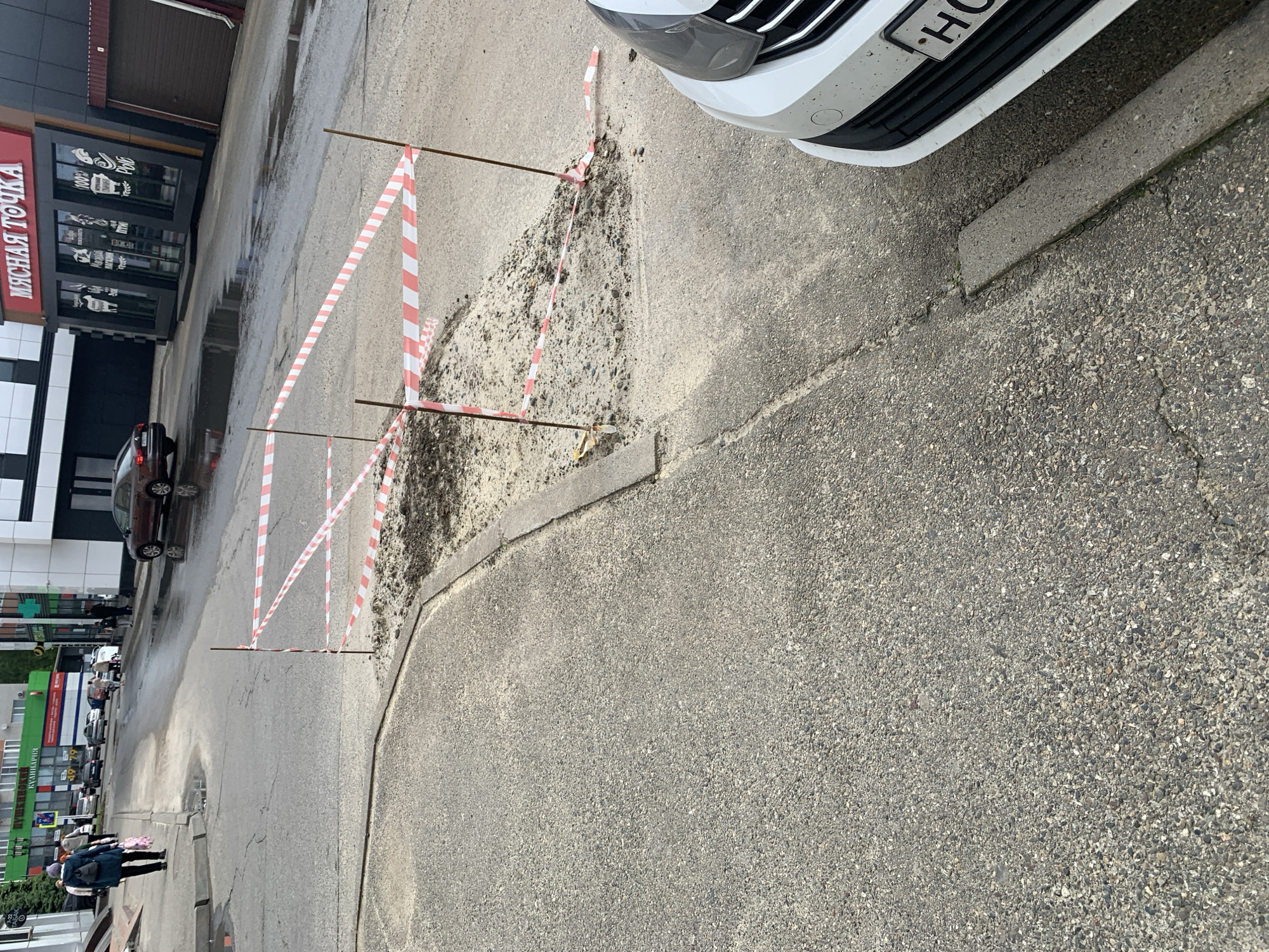 The image captures an urban street scene in Stavropol, Russia, featuring a hazardous road condition sectioned off by caution tape.
In the foreground and midground, a substantial pothole or area of damaged pavement is visible, filled with loose gravel and dirt. This area is cordoned off by red and white striped warning tape, which is stretched between several thin metal rods or stakes embedded in the ground. To the right, the front bumper and grille of a white car are partially visible, with the letter "H" clear on its license plate. A concrete curb runs parallel to the car, separating the main road from a slightly raised paved area. The asphalt surface of the road appears wet in various spots, suggesting recent rain or damp conditions.
In the background to the left, several buildings line the street. A prominent building features a red sign with white Cyrillic text that reads "МЯСНАЯ ТОЧКА" (Myasnaya Tochka), meaning "Meat Point" or "Meat Shop." Below this, smaller text like "100%" and "Свежее Мясо" (Fresh Meat) can be discerned. Adjacent to it, another building has a modern black facade with white checkered patterns and a dark roller shutter. Further down the street, a dark-colored car is parked, and a green sign for "ВЕТЕРИНАРИЯ" (Veterinariya - Veterinary) is visible on another building.
Several pedestrians are seen walking on the sidewalk in the distance on the left side of the frame. The overall impression is that of a bustling street on an overcast day, with immediate attention drawn to the marked-off road damage.