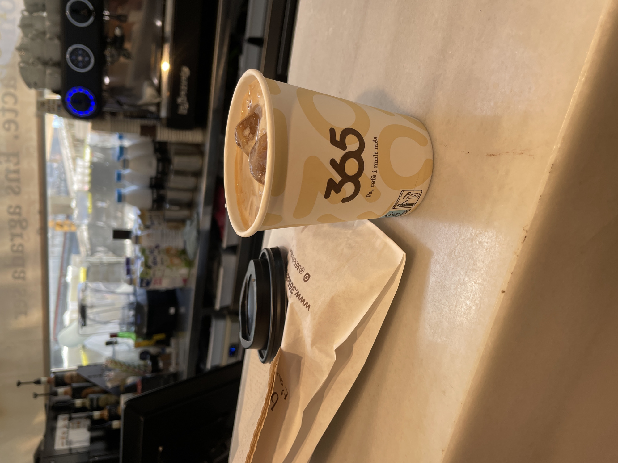 The image shows a paper cup of iced coffee on a counter, with a black plastic lid resting on a paper bag next to it. The cup has the "365" logo and the phrase "Pa, cafè i molt més," indicating it is from the 365.cafe chain, a popular coffee shop in Barcelona, Spain. The logo on the cup also includes a recycling symbol advising to recycle in the blue bin ("RECICLA AL AZUL" is Spanish for "Recycle in the blue bin").
In the blurred background, there's a coffee machine and a display of other beverages and snacks. The background suggests the setting is inside a café or shop. The counter is a light beige color.