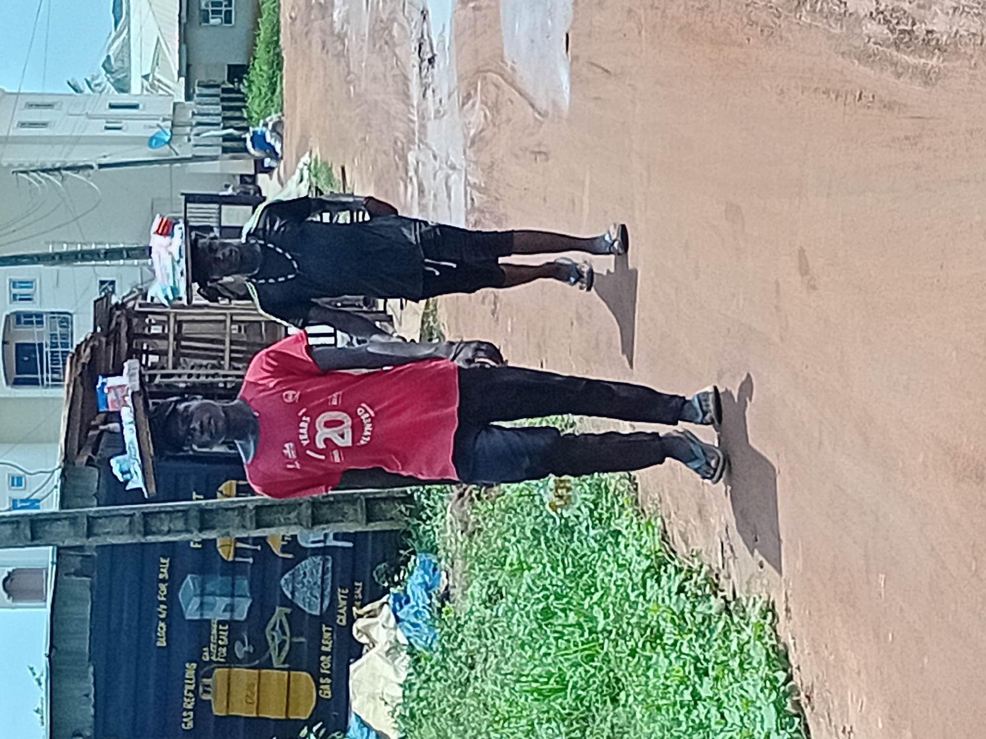 The image depicts two young men standing in Ikorodu, Nigeria, along a dirt road. One man wears a red shirt with "20 Years" printed on it and black pants. The second man is dressed in black shorts and a black shirt, sporting a necklace. Behind them, a sign advertises "Gas Refilling," "Block 4/9 For Sale," "Gas For Rent," and "Granite Sale," suggesting a commercial area. Lush green vegetation grows between the sign and the road. In the background, there is a building complex, indicating an urban setting.