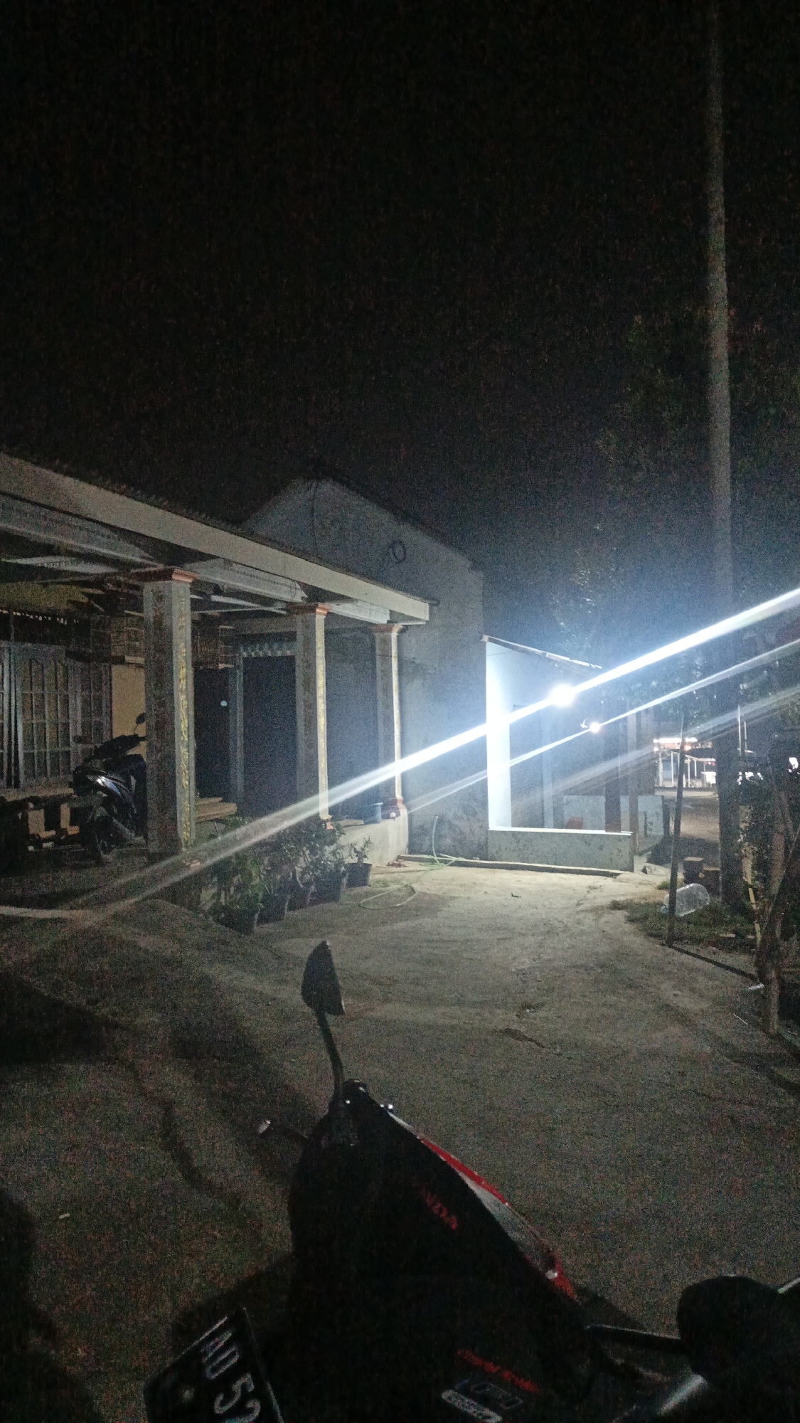 The image depicts an outdoor scene at night, likely in a residential area of Sambi, Indonesia. A motorcycle is prominently featured in the foreground, with its handlebars and part of the seat visible. The license plate of the motorcycle, reading "AD 52", is partially visible.
In the mid-ground, there is a building with a covered porch supported by several pillars. The building appears to be made of concrete, and a bright light source is illuminating the front of it, creating strong lens flares across the image. There are several potted plants lined up along the porch. A dark-colored motorcycle is parked under the porch to the left.
The ground is paved with concrete or asphalt, and there are shadows cast by the lighting. Beyond the building, trees and some distant structures are faintly visible in the darkness. A tall, slender pole stands to the right of the image. The overall impression is of a quiet, possibly rural or suburban, setting at night.