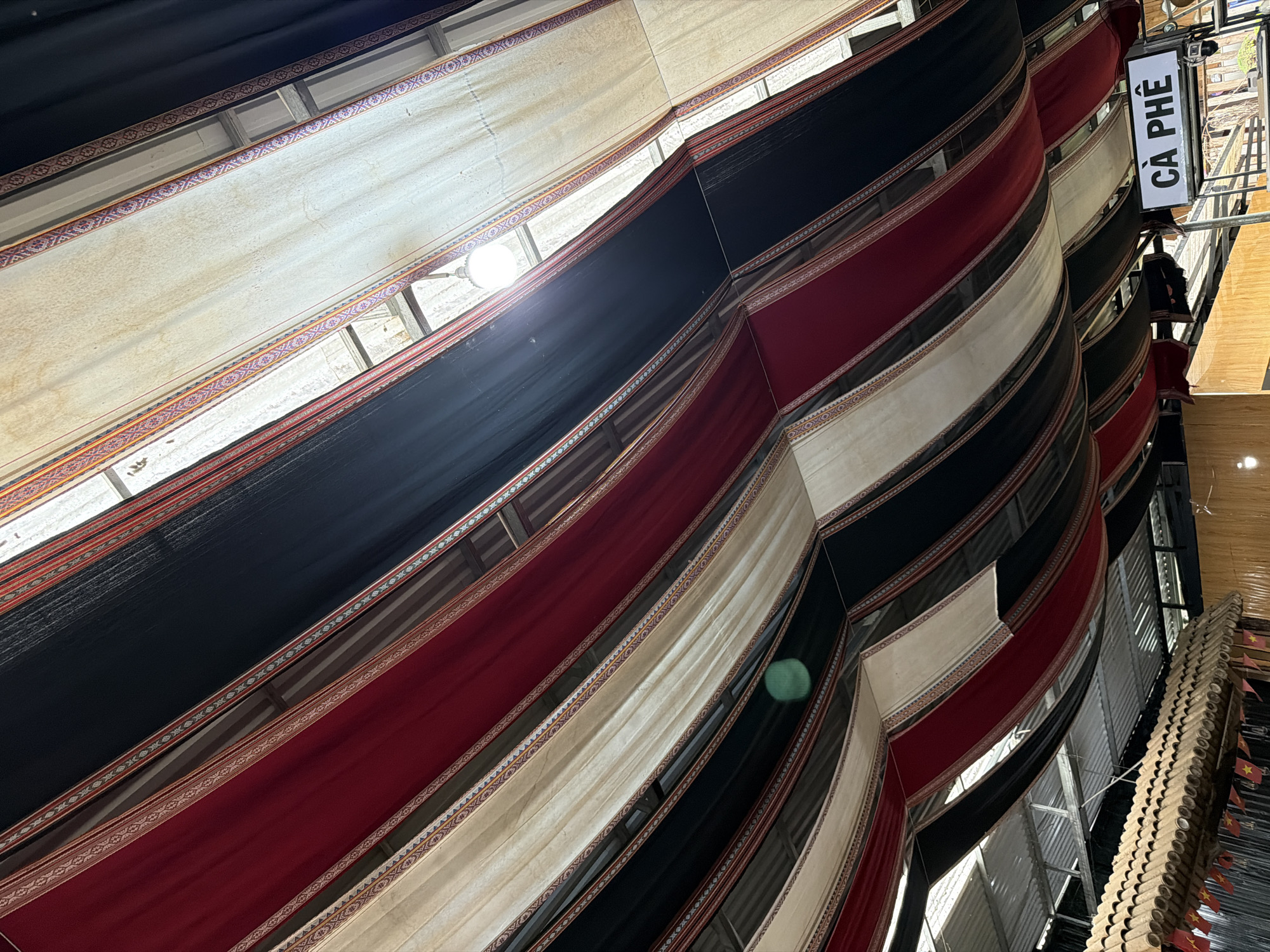 The image presents an upward-angled view of a decorative ceiling or canopy within an indoor or covered space, located in Măng Đen, Vietnam. Large panels of fabric, predominantly black, deep red, and off-white/cream, are arranged in flowing, layered sections overhead. Each fabric panel is edged with a distinct decorative trim featuring intricate, colorful geometric patterns, characteristic of traditional woven designs.
On the right side of the frame, a rectangular sign with the Vietnamese word "CÀ PHÊ" (coffee) is prominently displayed in black capital letters on a white background. Below this sign, parts of structural beams and what appears to be a corrugated metal roof or ceiling are visible. A bright light source, likely an artificial light fixture or sunlight, creates a glare in the upper left, and a soft green lens flare is present towards the center-right.
Further to the right, a unique vertical structure composed of numerous small, light-colored cylindrical elements, possibly bamboo or wood, is visible. This structure is adorned with multiple miniature Vietnamese national flags, each featuring a red field with a yellow star. The scene suggests a culturally rich environment, possibly a café or a cultural display area, with elements reflecting Vietnamese heritage.