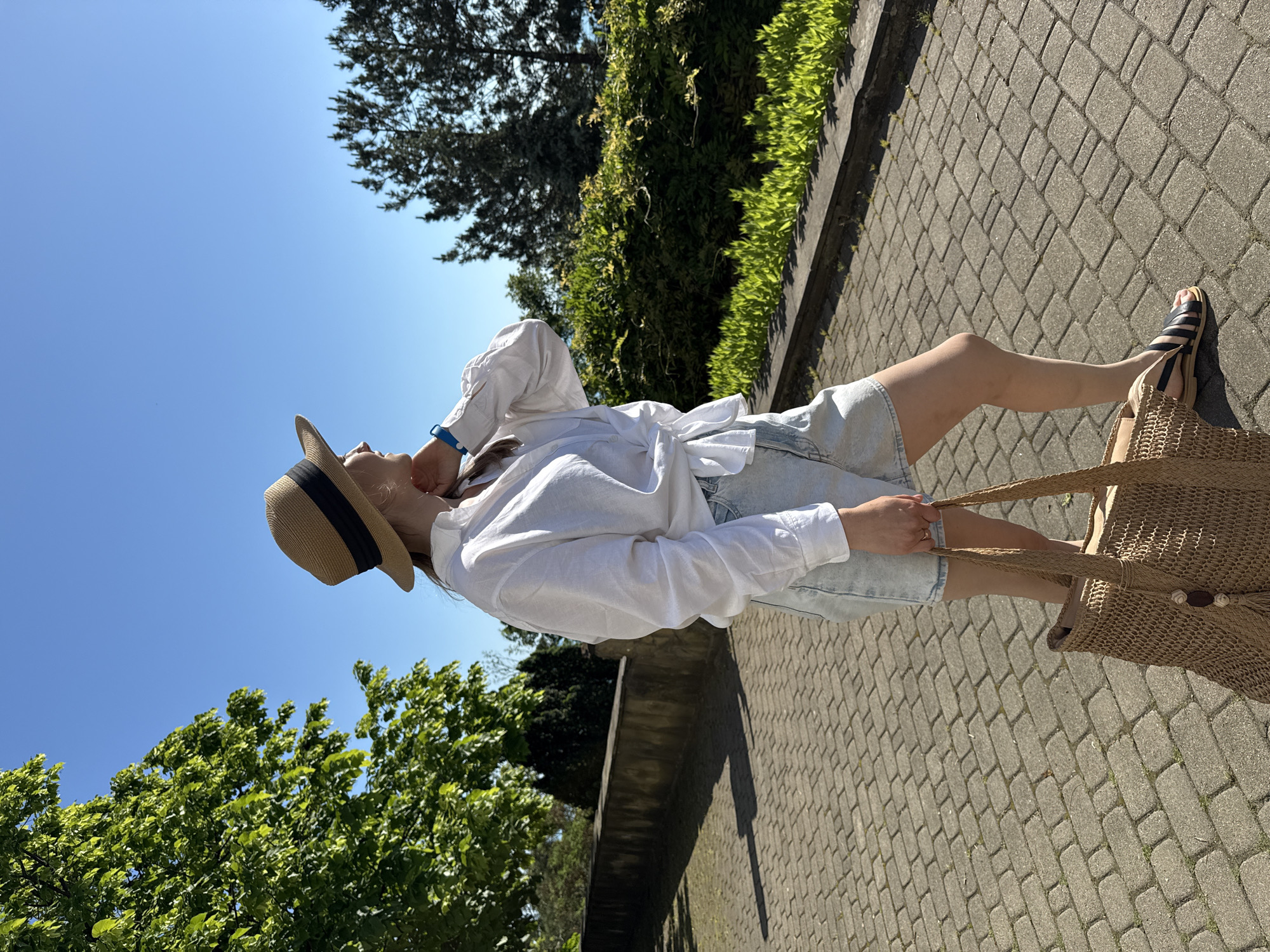 The image features a person standing outdoors on a bright, sunny day in Nebug, Russia. The individual, likely female, is dressed in a casual summer outfit. They are wearing a loose-fitting white long-sleeved shirt, light-wash denim shorts, and black and white striped flat sandals. A straw hat with a dark band adorns their head, and a blue wristband is visible on their left wrist. They are holding a large, light brown woven straw bag by its strap in their right hand.
The person is captured looking upwards, with their head tilted slightly back and their left hand raised towards their neck or hat, suggesting a moment of contemplation or enjoyment of the clear sky. They are standing on a paved pathway constructed from interlocking grey pavers, which exhibits sharp shadows indicating strong sunlight. The setting is rich with greenery; lush trees and bushes line the path, particularly visible on the left side of the frame. Above, the sky is a clear, vibrant blue, devoid of clouds.
The overall scene evokes a relaxed, summery atmosphere. The distinct shadows and bright sky point to a daytime setting, likely mid-morning or afternoon. There is no visible text in the image.