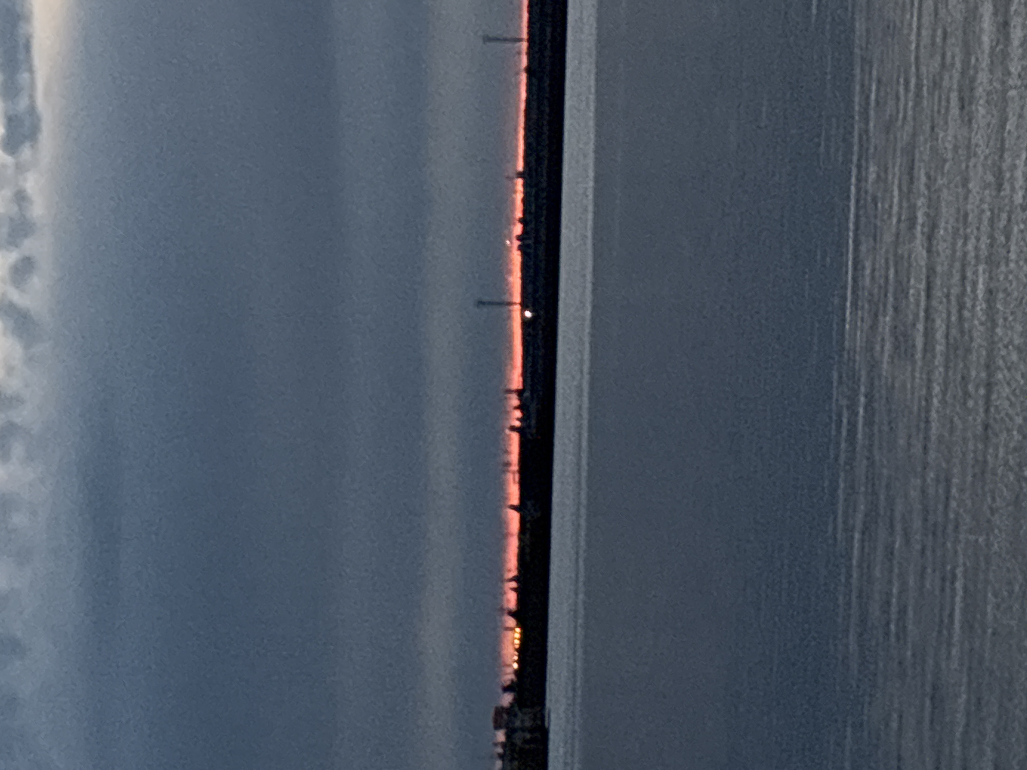 The original image, when viewed horizontally, depicts a tranquil twilight scene over a large body of water, likely the Votkinsk Reservoir near Chaikovsky, Russia. The composition is divided by a subtle horizon line that stretches across the middle.
**Subjects:** The upper portion of the image shows a dark, overcast sky, dominated by muted shades of grey and blue, hinting at the lingering light of dawn or dusk. The lower portion reveals a calm expanse of water, reflecting the subdued sky and appearing dark and deep.
**Activity or Event:** The image captures a peaceful moment of transition between day and night, highlighting the natural beauty of the landscape at low light. There are no visible people or discernible activities.
**Notable Details:** A thin, dark line delineates the horizon, suggesting a distant landmass or an artificial structure like a causeway or bridge. Along this distant line, several small, glowing points of light are visible, which could be streetlights or structures on the far bank. A faint, reddish-orange glow illuminates the sky directly above this horizon line, indicative of either a sunset or sunrise breaking through the clouds. The water surface exhibits gentle texture, implying very still conditions. The overall mood is quiet and atmospheric.
**Visible Text:** There is no visible text in the image.