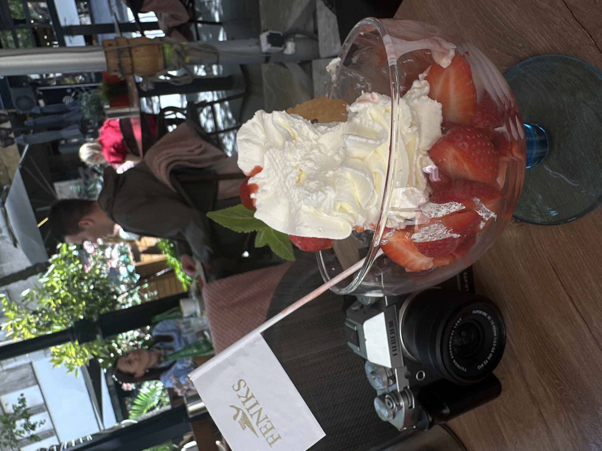 The image depicts a table in an outdoor cafe, likely in Belgrade, Serbia, given the presence of a "FENIKS" flag in the foreground, a chain with multiple locations in the city. A dessert is prominently featured, served in a stemmed glass, containing sliced strawberries, whipped cream, and what appears to be a biscuit or cookie. To the right of the dessert is a Fujifilm camera with a lens. In the background, there are blurred figures of people, including a woman in a blue shirt and other people. The environment includes trees, possibly part of the café's outdoor setting, along with tables, chairs, and overhead structures. The overall composition suggests a casual outdoor dining experience.