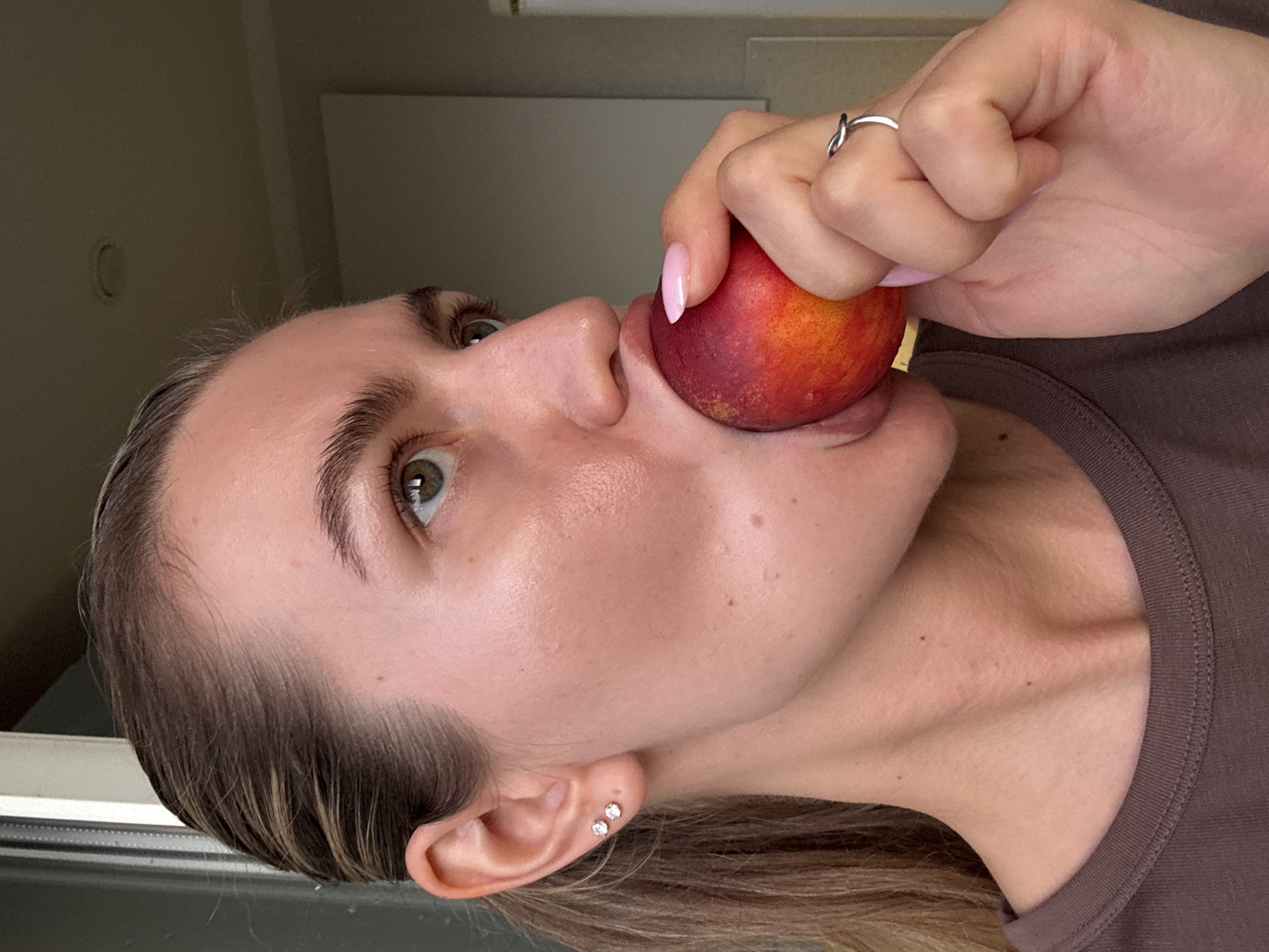 A close-up, profile shot shows a woman's face and upper body as she holds a peach to her mouth. The woman has light skin, brown hair pulled back in a ponytail, and green eyes. She is wearing a brown t-shirt, small stud earrings, and a silver ring. Her fingernails are painted pink. The background is indistinct, but appears to be indoors, possibly near a window. The lighting suggests it is daytime.