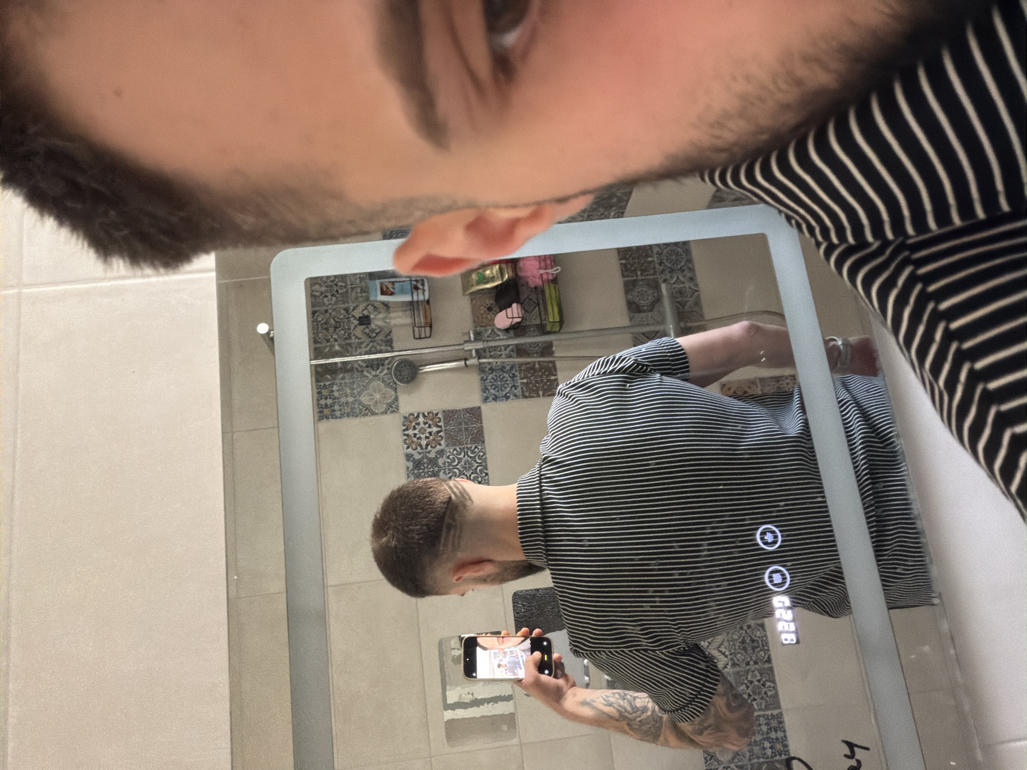 A man takes a selfie in a mirror in a bathroom. The man is wearing a black and white striped shirt and has tattoos on his right arm. He is holding a smartphone in his left hand. The bathroom has patterned tiles on the wall, a shower, and shelves. The mirror has touch screen icons that can be seen. The location is Yoakim Gruevo, Bulgaria.