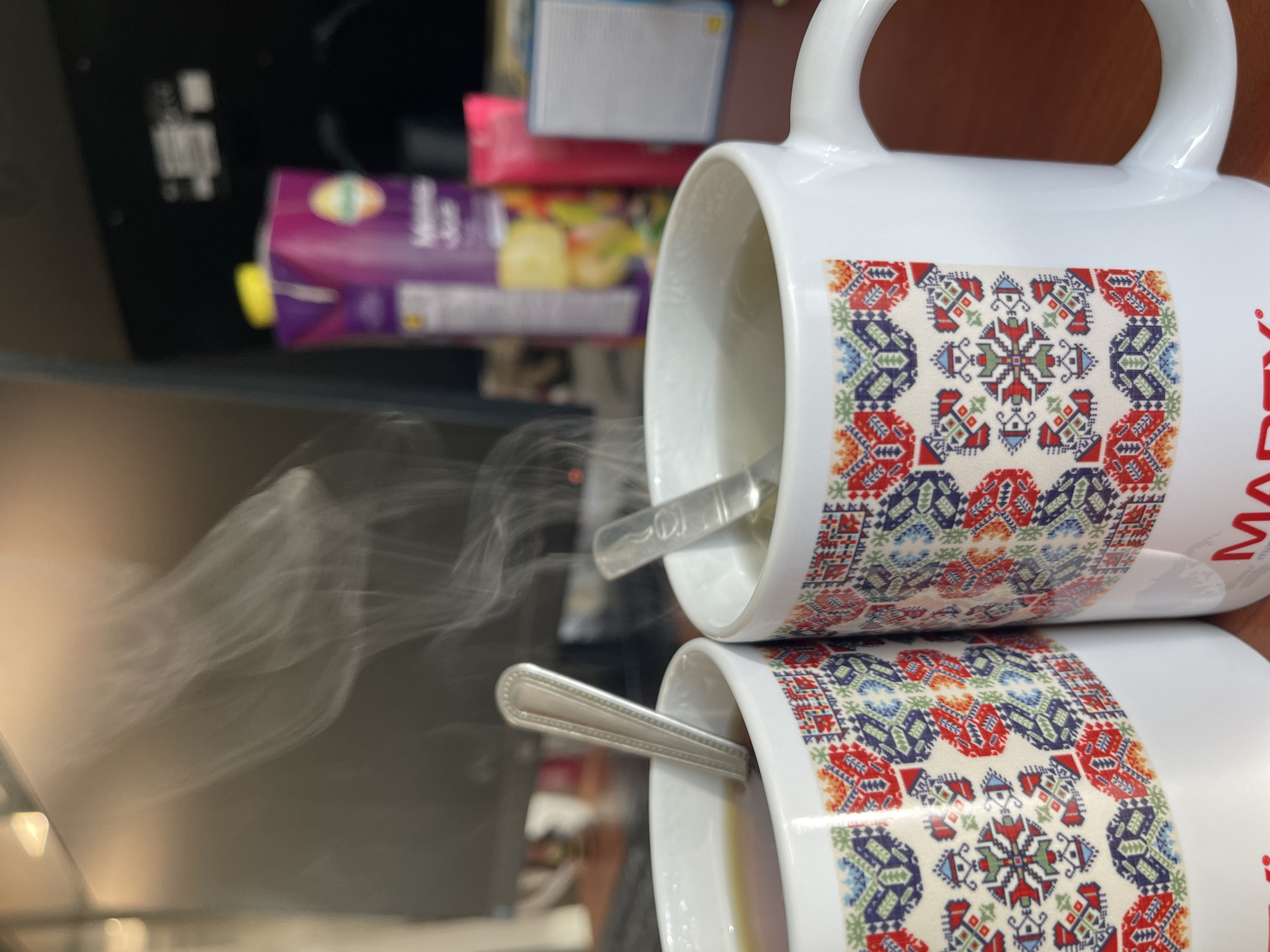 The image shows two stacked white mugs, each with a handle and a design featuring a pattern of red, blue, and green geometric shapes. Steam is rising from the mugs, suggesting they contain hot beverages and indicating the location is Sofia, Bulgaria. Silver spoons are placed inside each mug. The foreground is blurred, while the background reveals various objects, including a purple juice box and other items stacked behind the mugs. Part of the word "MARTEX" is visible on one of the mugs.