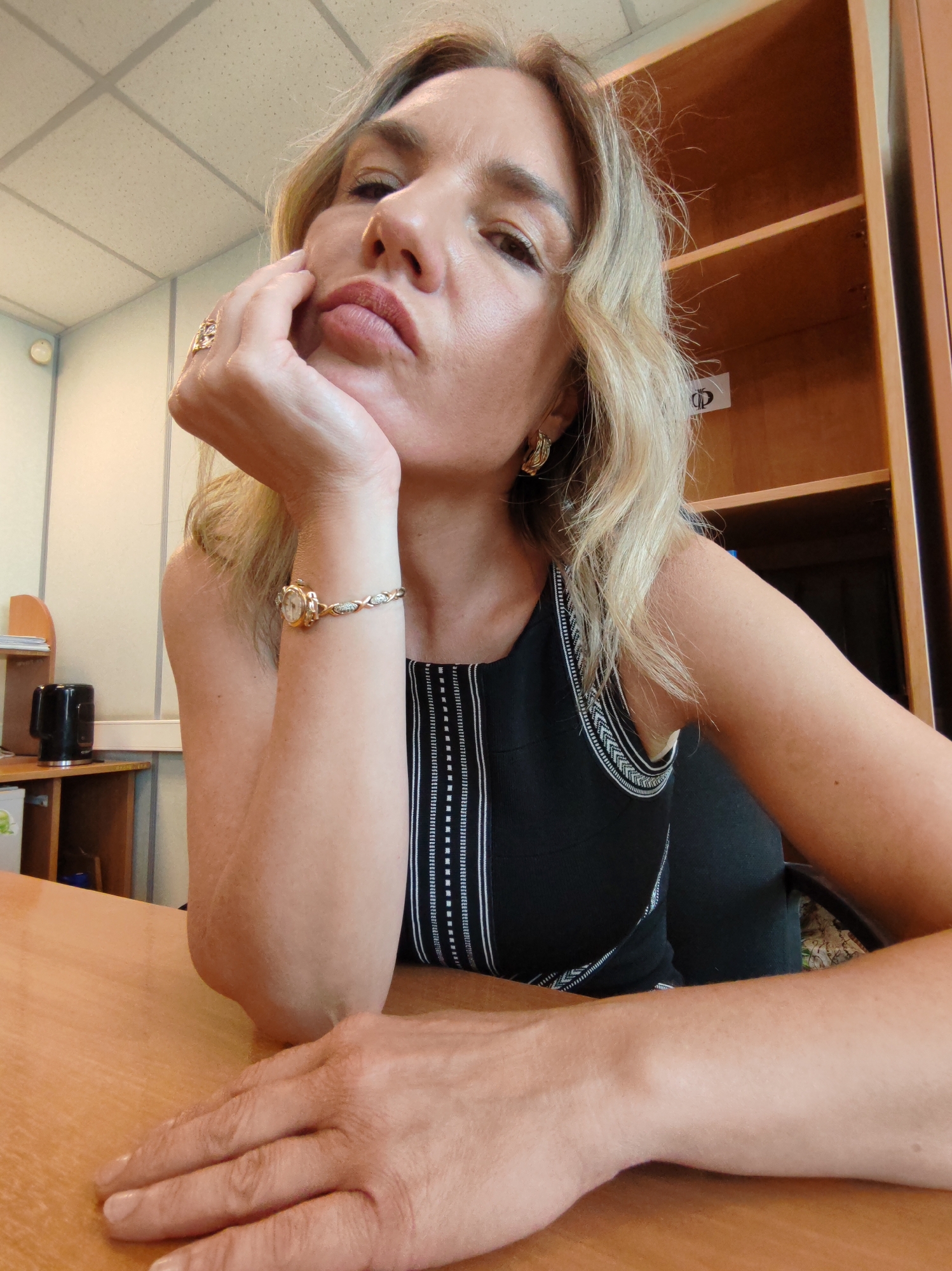 A woman is sitting at a wooden desk in what appears to be an office setting. She has blonde hair and is wearing a sleeveless black top. She is resting her chin on her hand, and her facial expression appears contemplative. She is wearing a watch and a ring. The background shows office furniture, including a shelf unit and other desk items. The location is Unknown, Unknown.