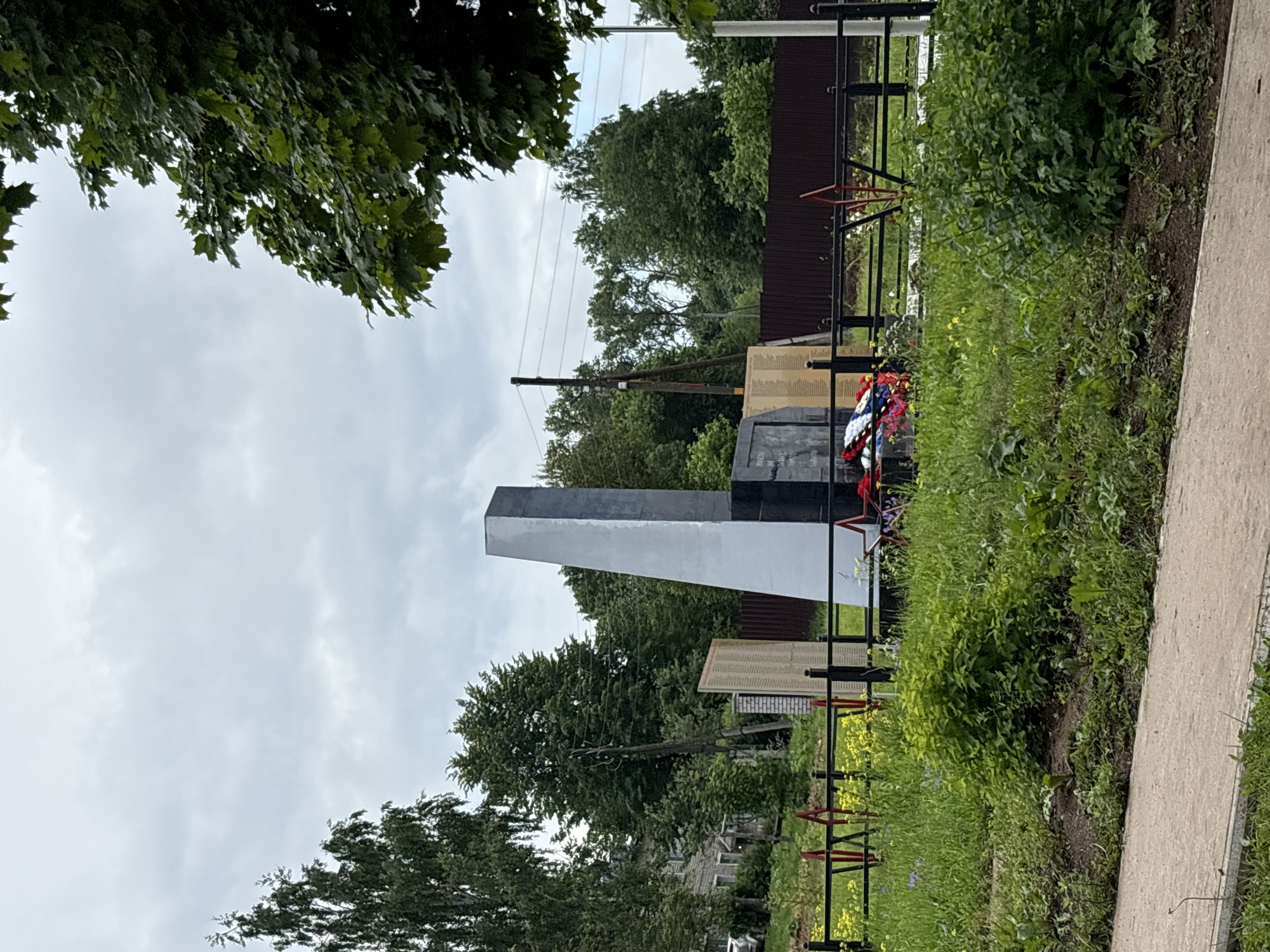 The image depicts a war memorial located in Kstino, Russia. The memorial consists of a tall, obelisk-like structure that is partially grey and partially black. In front of the obelisk, there are wreaths of red, white, and blue flowers. To the left of the obelisk, there is a flat monument or plaque with inscriptions, partially obscured by the main memorial. Behind the memorial, a dark brown fence runs horizontally across the scene, with a few utility poles visible in the distance. The surrounding area is overgrown with green grass and some yellow wildflowers. Trees with dense foliage form a backdrop to the memorial. A paved walkway runs along the bottom of the image, in front of the grassy area. The sky is overcast and cloudy. No people are visible in the image.