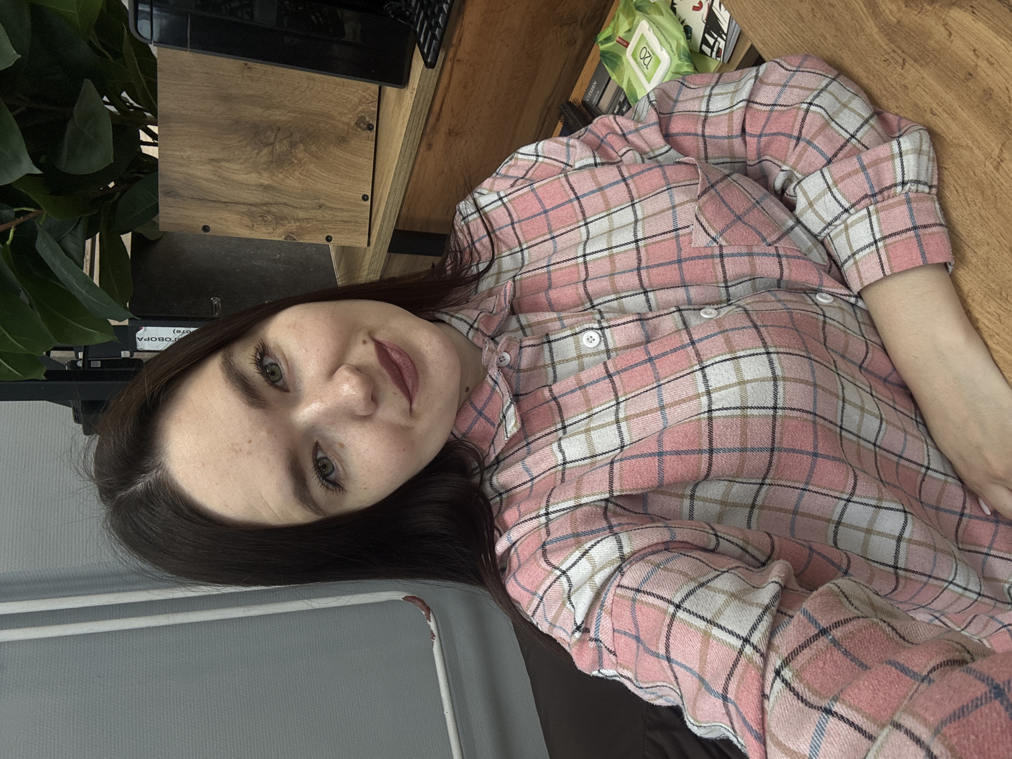 A woman with dark hair and light skin is wearing a pink and white plaid shirt. She is indoors with her head tilted to the left. A wooden desk or shelf is behind her, with a green plant and a dark object on top of it. A grey surface is visible in the lower left corner. The location cannot be confirmed as Chaikovsky, Russia based on the image.