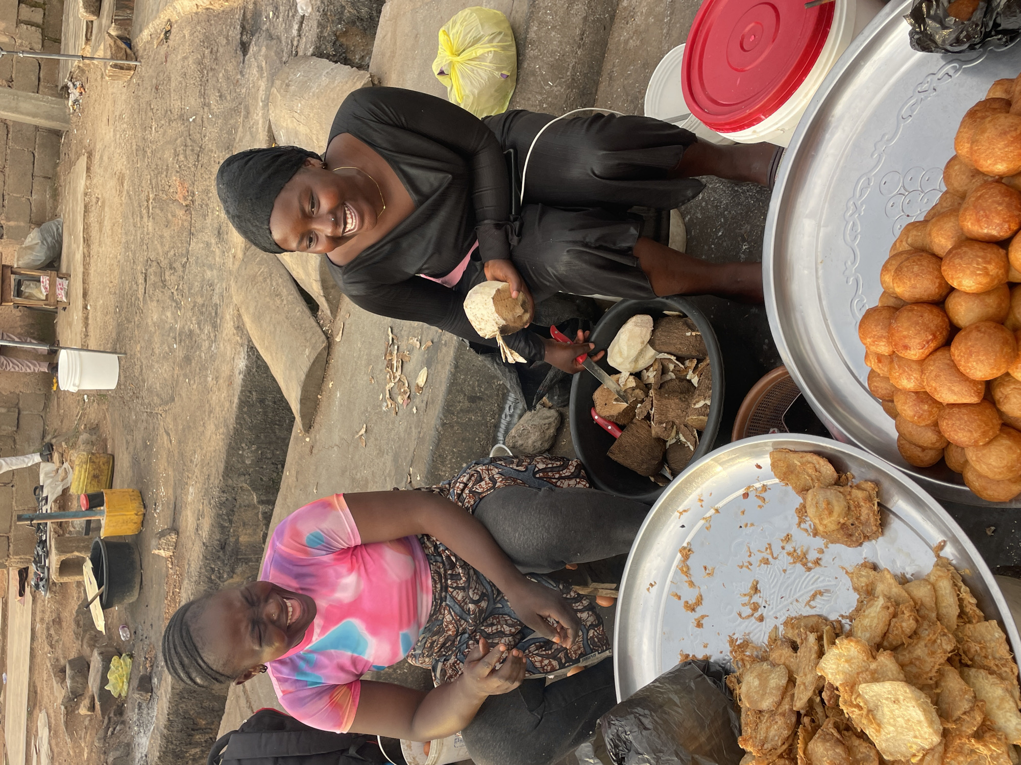 Two Nigerian women are captured in a candid moment of laughter in Jos, Nigeria. The woman on the left, wearing a tie-dye t-shirt and a patterned skirt, is mid-laugh with her eyes closed. Her hair is styled in braids. Next to her, the woman on the right, dressed in a black headscarf and a black dress, is also laughing heartily. She is holding a piece of coconut and a knife, suggesting she might be preparing food.
In the foreground, two large metal trays are filled with food. The tray on the left holds an assortment of fried snacks, appearing crispy and golden brown. The tray on the right is piled high with round, puffed fried dough balls, also golden brown. Behind the women, several black buckets contain more food items and tools, including a large black bucket filled with cut coconuts and a knife. A yellow plastic container and a black plastic bag are also visible. The ground is dirt and concrete, with some scattered debris, typical of an outdoor market setting in Jos. The overall scene depicts a vibrant and lively market atmosphere, with the women's joyful expressions adding a personal touch.