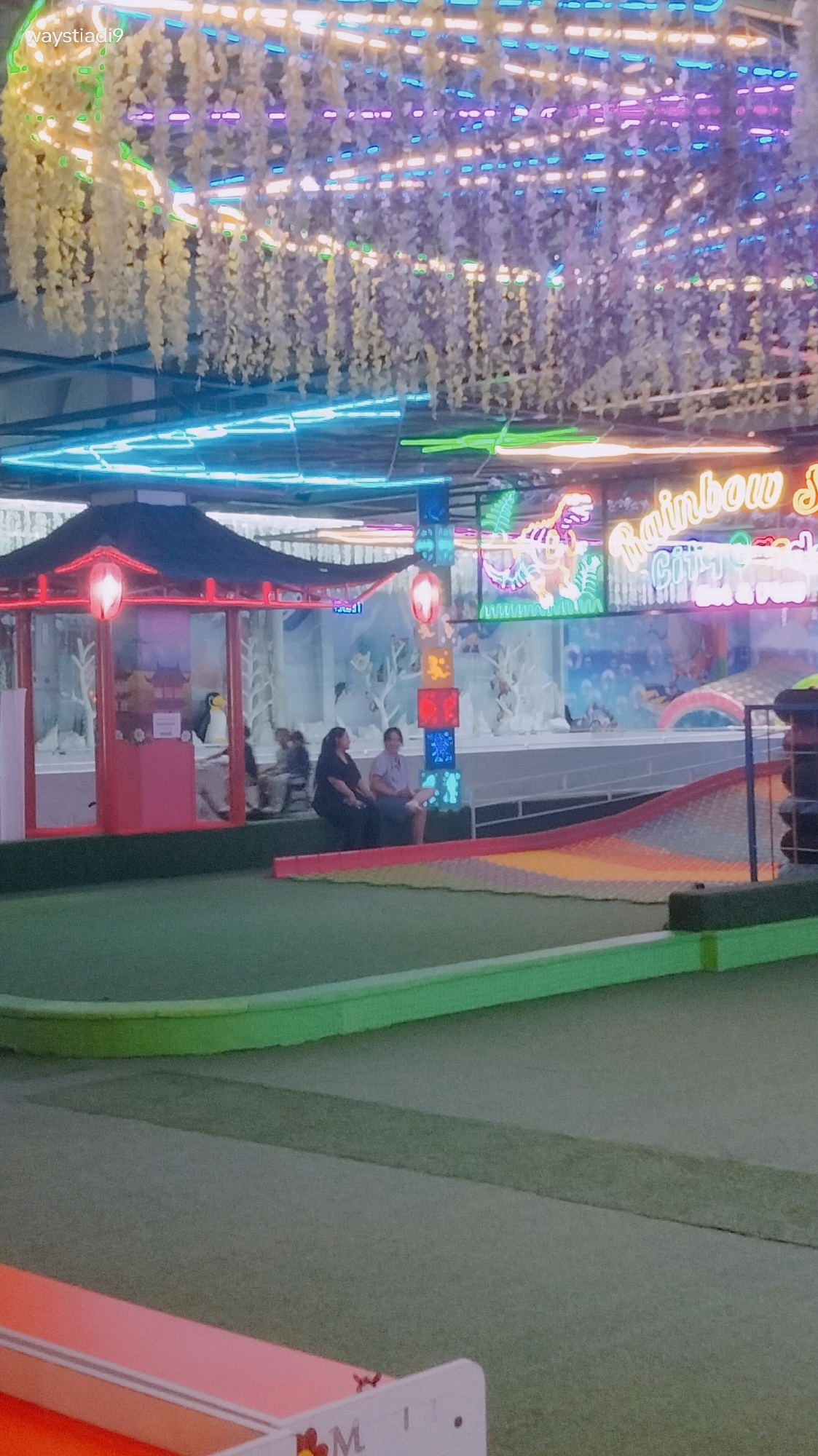 The image depicts an indoor entertainment area, likely a theme park or arcade, with vibrant neon lights and decorative elements. A prominent sign reads "Rainbow City" in illuminated lettering, suggesting a playful and colorful atmosphere. Artificial greenery, resembling vines, hangs from the ceiling, interspersed with strings of lights in purple, blue, and yellow.
In the midground, a small, red and black traditional-looking building with a dark roof is visible. Two individuals are seated on a bench in front of this structure. To the right of this building, there's a multi-colored, soft-surfaced ramp, possibly part of a playground or attraction. Behind the building and ramp, there's a backdrop with what appears to be icy or snowy scenery, complete with illuminated figures and abstract shapes.
The foreground shows a green, artificial turf surface, possibly a mini-golf course, with a light-colored border. Part of a pink structure with some text and a cartoonish character is visible at the very bottom of the frame. The overall impression is that of a lively and modern recreational space. Based on the provided information, this scene is located in Bandung City, Indonesia.