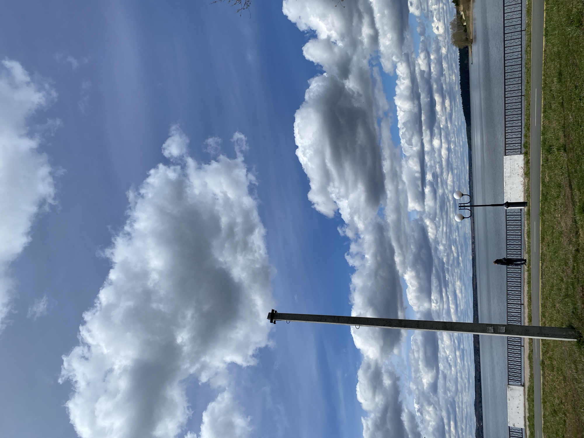 The image shows a partially cloudy sky above a lakeside scene in Votkinsk, Russia. The sky is mostly blue with numerous puffy white clouds. A concrete utility pole stands in the foreground. Adjacent to the lake is a paved path, delineated by a metal railing with a distinctive wave pattern. A person stands near a lamppost along the path. Grass lines the edge of the path on the far side. The lake is visible beyond the path and railing, and a line of trees is visible on the opposite shore.
