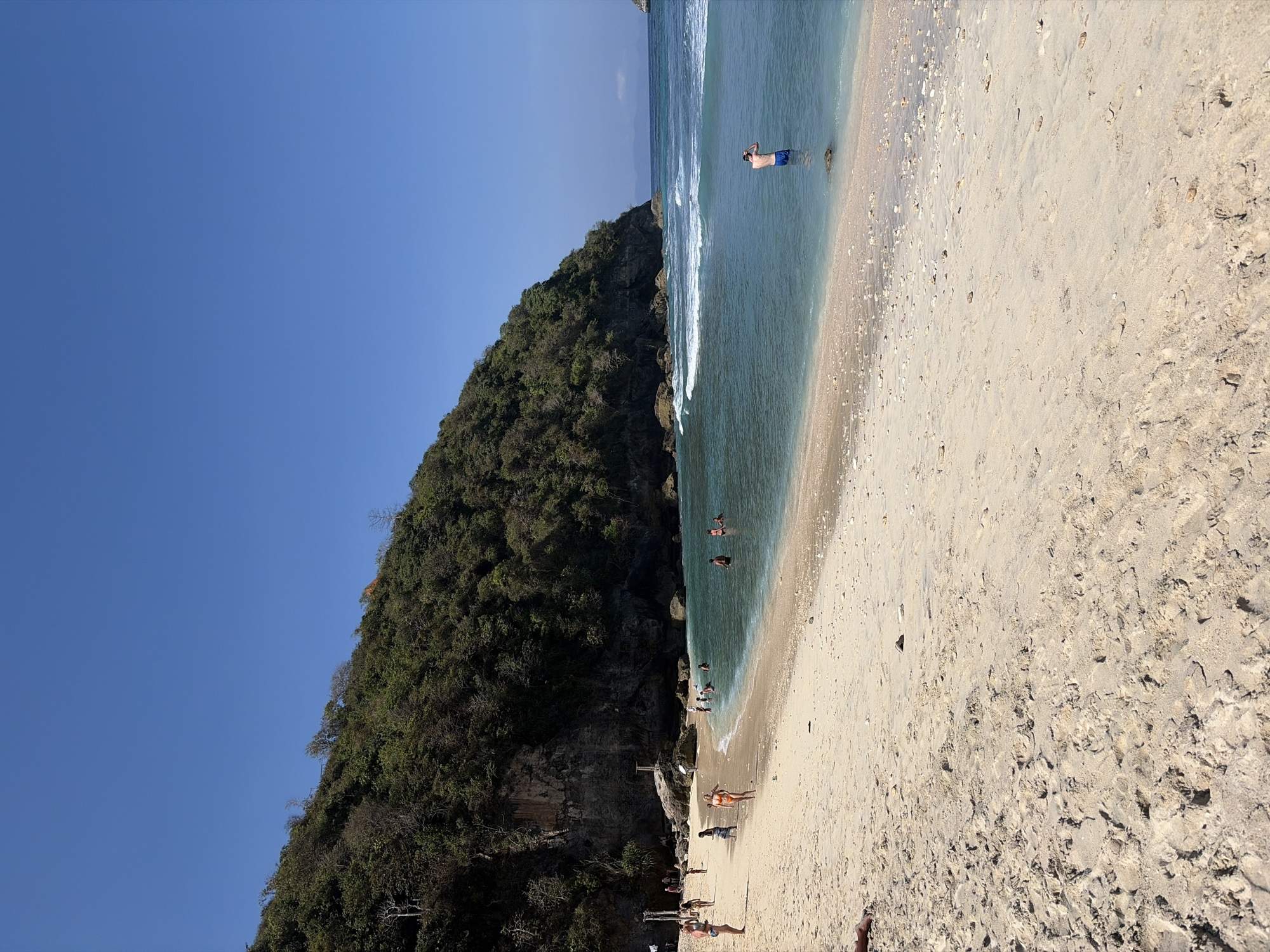 The image depicts a crowded beach in Nusa Penida, Indonesia, on a sunny day. The foreground is dominated by light-colored sand with scattered shells and footprints. In the middle ground, several people are visible on the beach and wading in the turquoise water. Some individuals are swimming, while others stand in the shallow water. The background features a steep, tree-covered cliff face that curves into the sea, with waves breaking against the shore. The sky is clear and blue. A single man in blue shorts is standing in the water in the right-middle ground, facing away from the camera.