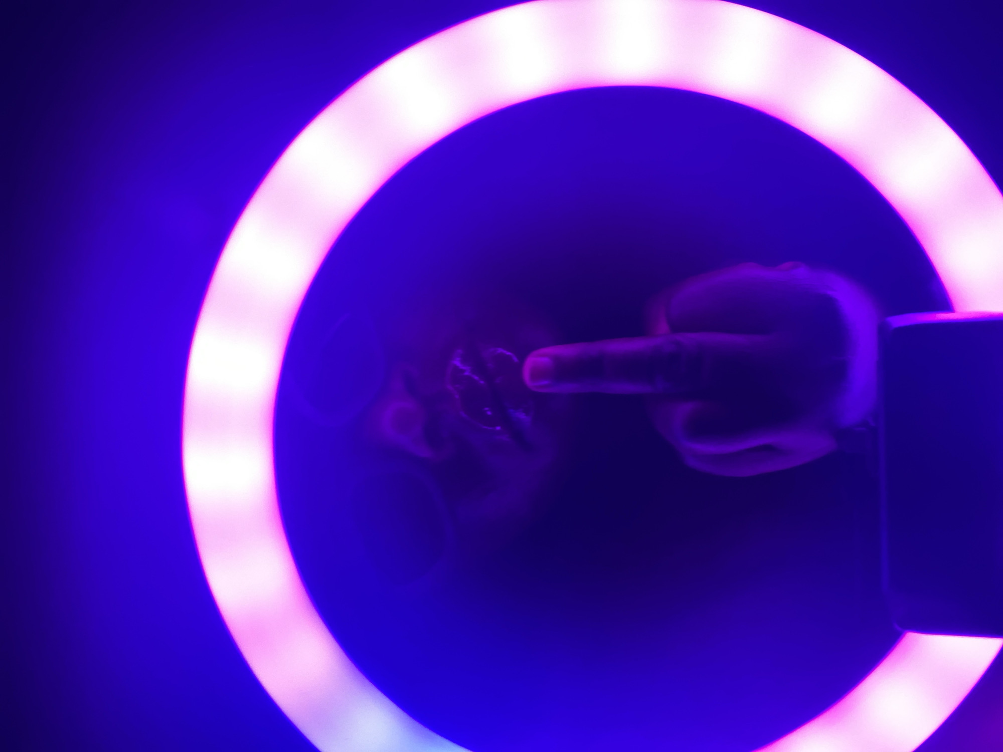 A low-light image captures a person's hand and a portion of their face, prominently framed and illuminated by a bright, glowing ring light. The primary colors in the scene are deep purple from the background and vibrant pink-white from the ring light.
The central focus is a human hand, specifically the middle finger, which is raised and extended directly in front of the camera, appearing to pass through the center of the ring light. The ring light itself is a circular source of illumination, casting a strong, soft glow that highlights the hand and parts of the person's face. Above and slightly to the left of the hand, the lower part of the person's face, including their chin and mouth area, is faintly visible, illuminated by the pink-white light. The individual's features are indistinct due to the lighting and slight blur.
To the right side of the ring light, a dark, rectangular object, possibly a smartphone or recording device, is partially visible, suggesting the image might be a selfie or a video recording in progress. The background is uniformly dark and out of focus, emphasizing the illuminated ring and the subject within it. The raised middle finger gesture typically conveys defiance, disrespect, or strong disapproval. The setting appears to be an indoor or controlled environment with artificial lighting, likely at night or in a darkened room, potentially in Maigizo, Nigeria. There is no visible text.