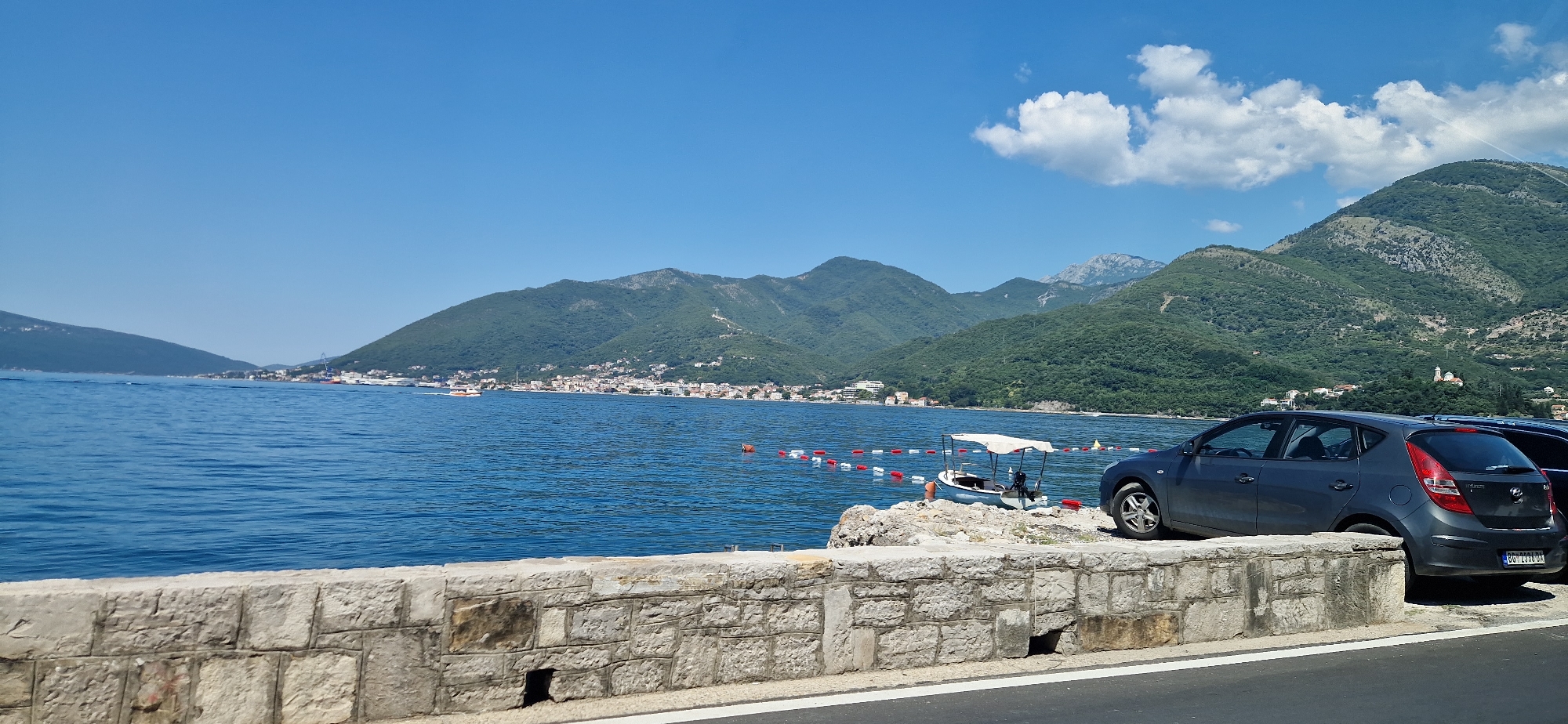 The image depicts a coastal scene in Donja Lastva, Montenegro, under a clear blue sky with scattered white clouds. In the foreground, a stone wall borders a paved area, likely a road or walkway, with a dark gray hatchback car parked to the right. The car has its rear bumper and taillights visible, and a license plate with characters including "BG 2338 DA". Beyond the wall and road, a calm blue body of water stretches towards a town nestled along the base of lush green mountains. A line of red and white buoys marks an area on the water, and a small white boat with a canopy is docked near the shore. The town exhibits a collection of buildings with red-tiled roofs. In the background, the imposing mountains are covered in dense vegetation, with some lighter gray rock formations visible on their slopes. A prominent white cross is situated high on one of the mountainsides. The overall atmosphere suggests a sunny day in a coastal Mediterranean town.