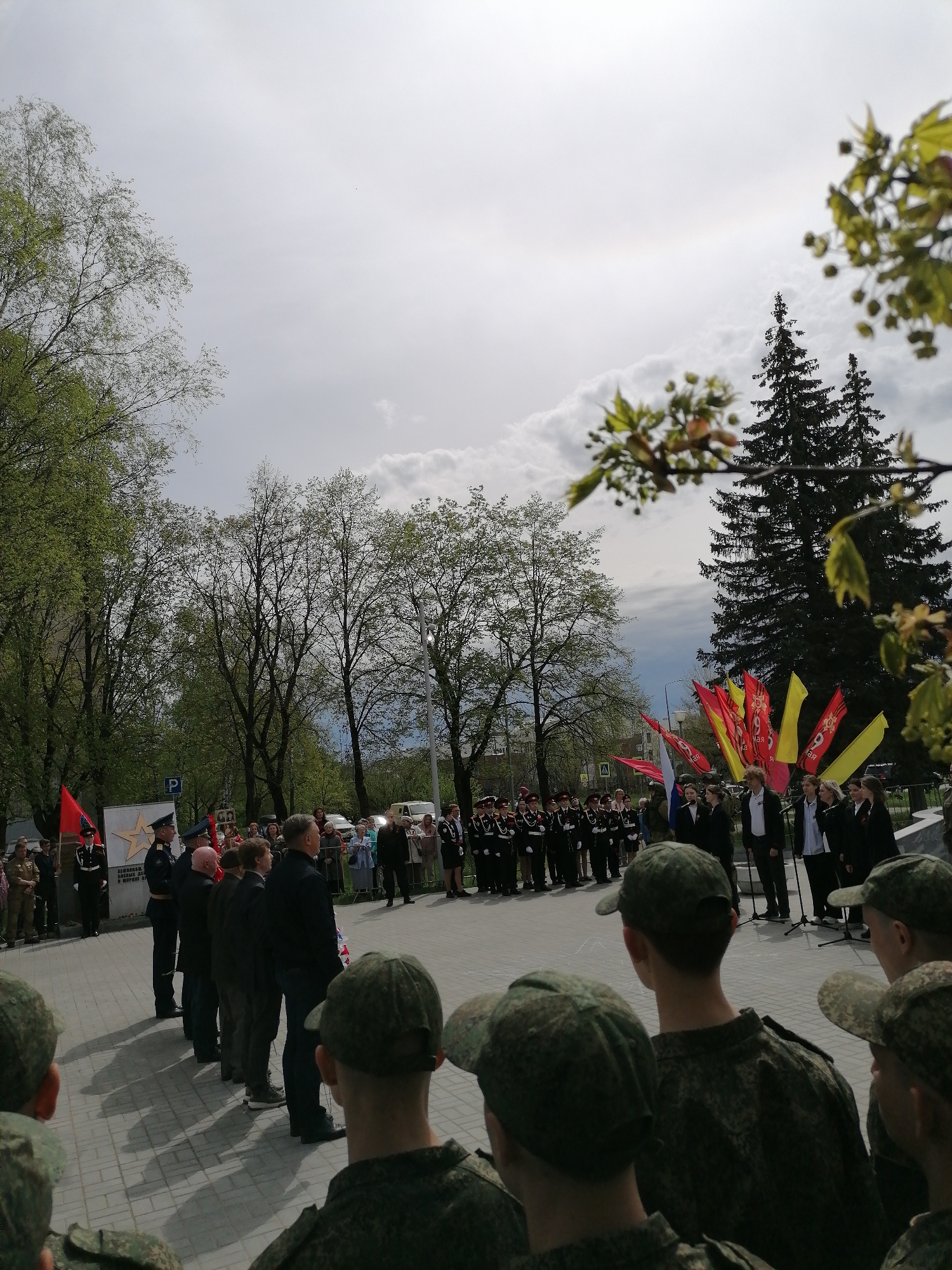 A group of people, including soldiers in camouflage uniforms and officers in dress uniforms, are gathered in a public area of Unknown, Unknown. They appear to be observing a ceremony or event, possibly a memorial, based on the presence of flags, microphones, and what seems to be a choir or singing group. There are trees surrounding the area, and the sky is overcast. A memorial or monument is visible on the left.