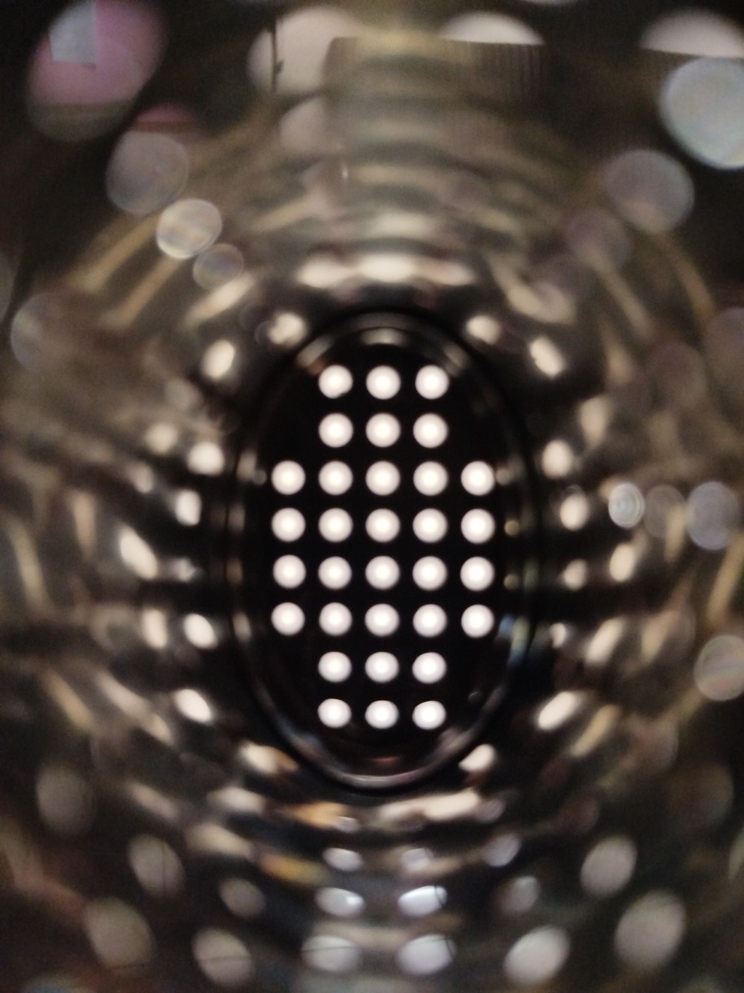 The image shows a close-up, interior view of a perforated metallic object, possibly a kitchen utensil like a grater or a colander, viewed from the inside looking out. The central focus is an oval-shaped opening with a grid of evenly spaced white circles, suggesting illuminated holes or the texture of the material. The surrounding interior surface is metallic, with blurred reflections and highlights creating a tunnel-like effect. There are no people, animals, or other objects visible. The image does not contain any specific geographical markers or clues that would allow for its location to be identified in Kremenchuk, Ukraine, or any other city.