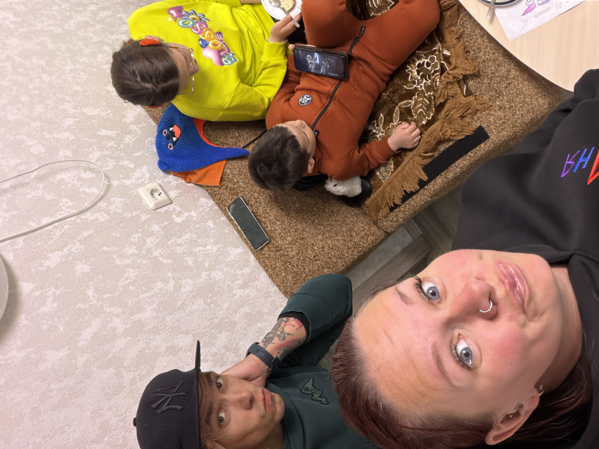 A high-angle shot shows a group of four people in an indoor environment.
Two children lie on a brown rug. One wears a bright yellow sweatshirt with colorful graphics, while the other wears an orange tracksuit. A smartphone rests on the child in the orange tracksuit. A drawing is visible on a table nearby.
Two adults complete the scene. One person, wearing a black hoodie, holds the camera for a selfie. Adjacent to them, another person wears a dark green sweatshirt and a black baseball cap, looking towards the camera. The background consists of a light-colored patterned floor, along with a white electrical outlet and a charging cable.
The location is Chaikovsky, Russia.